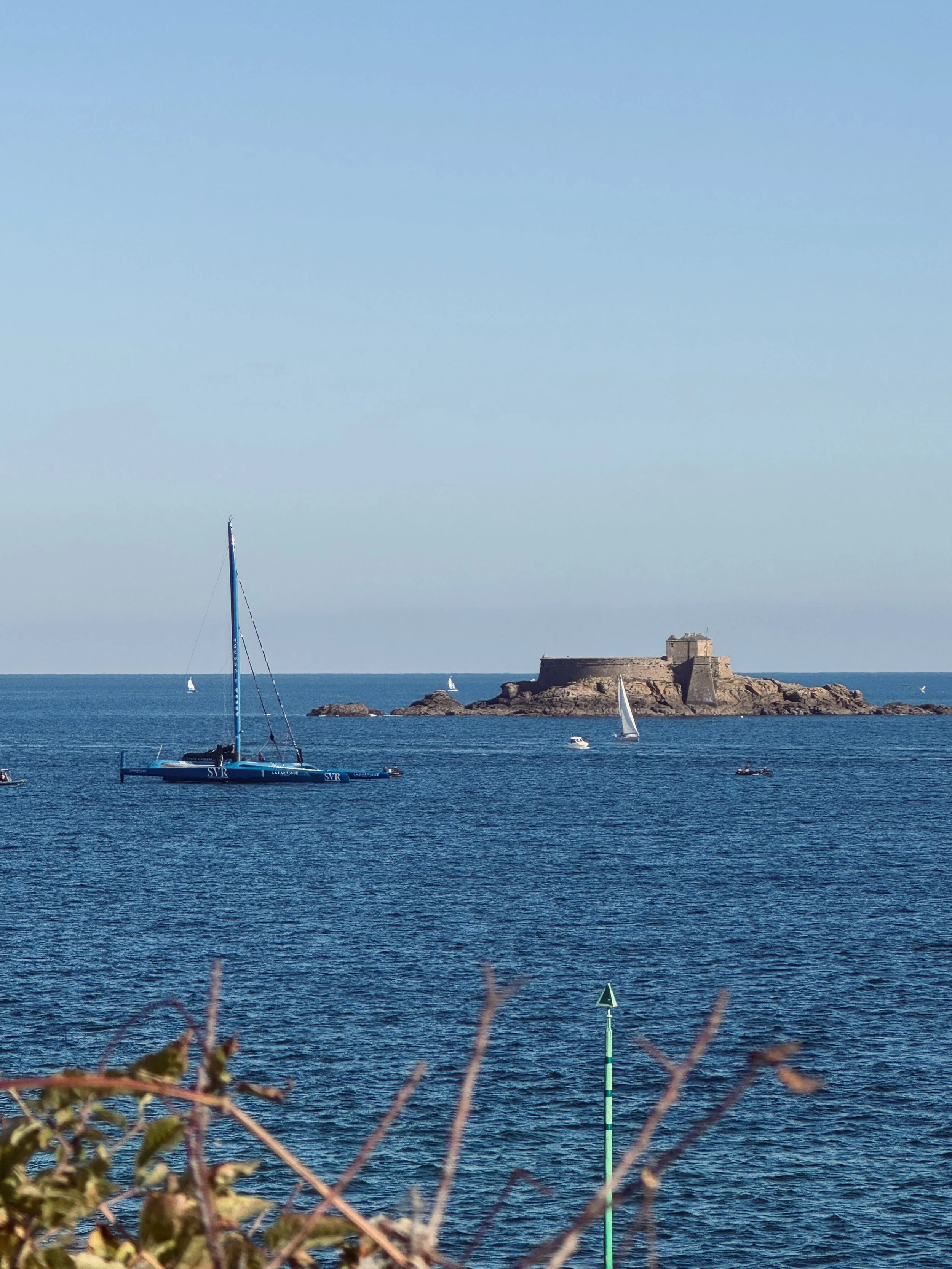 Voiliers naviguant devant un ancien fort côtier construit sur un îlot rocheux, avec une balise maritime au premier plan.
