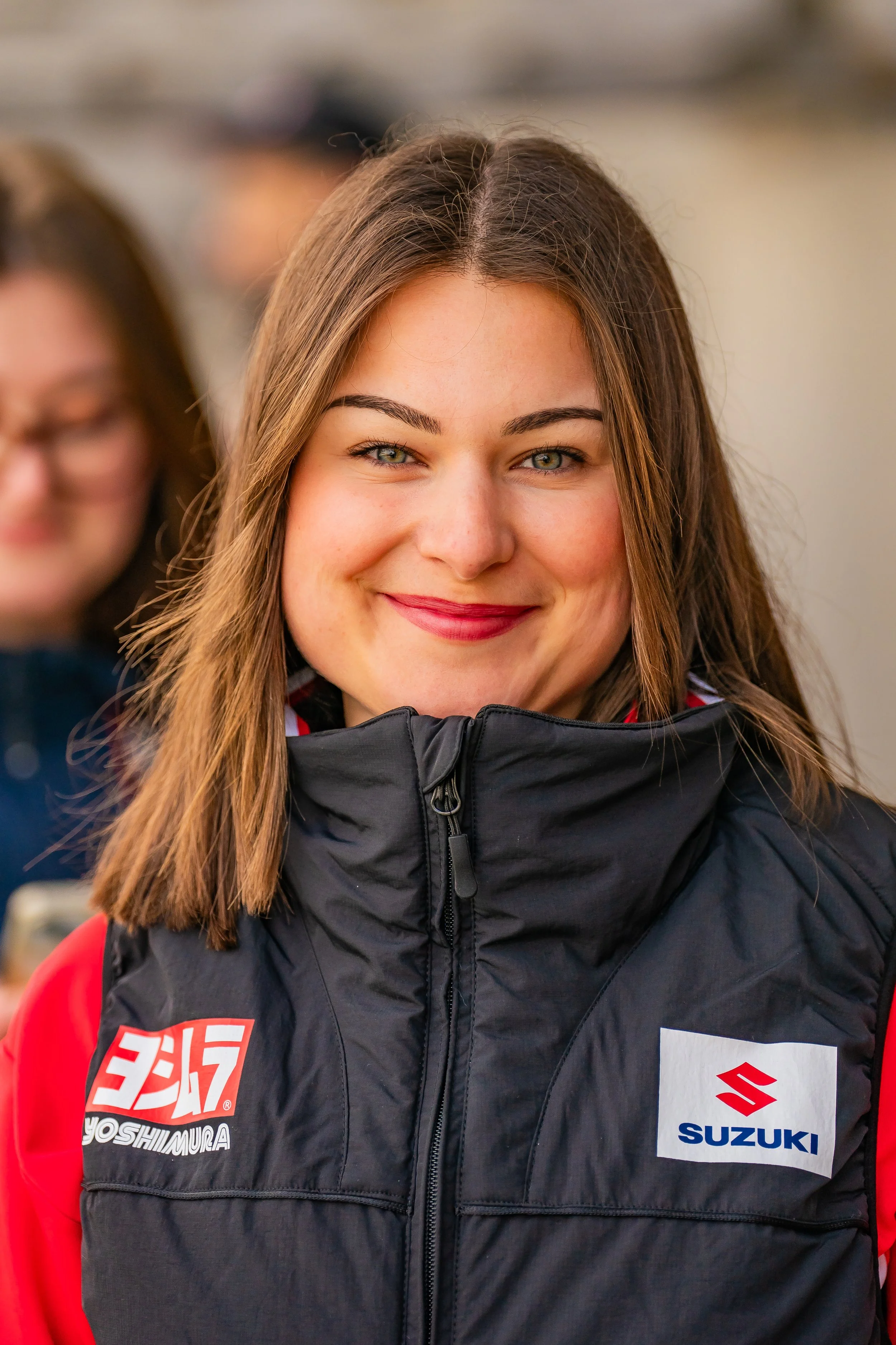 Portrait rapproché d’une femme souriante aux cheveux longs, portant une veste noire Suzuki avec logos Yoshimura et Suzuki.