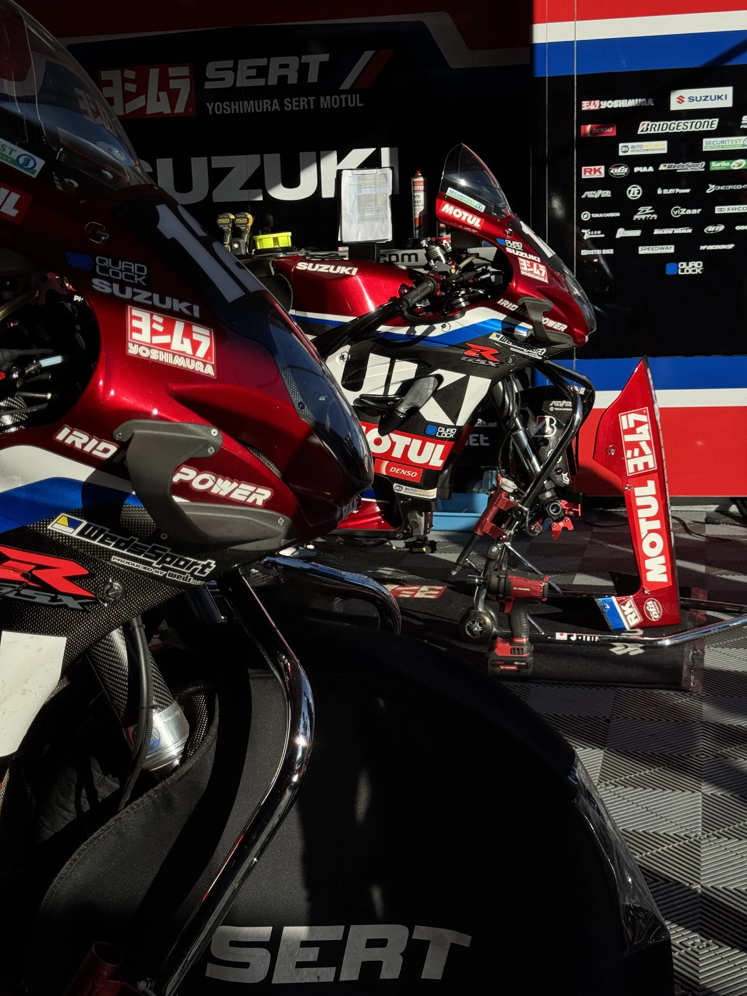 Moto de course Suzuki Yoshimura SERT Motul rouge et noire, entourée de logos de sponsors dans un paddock.