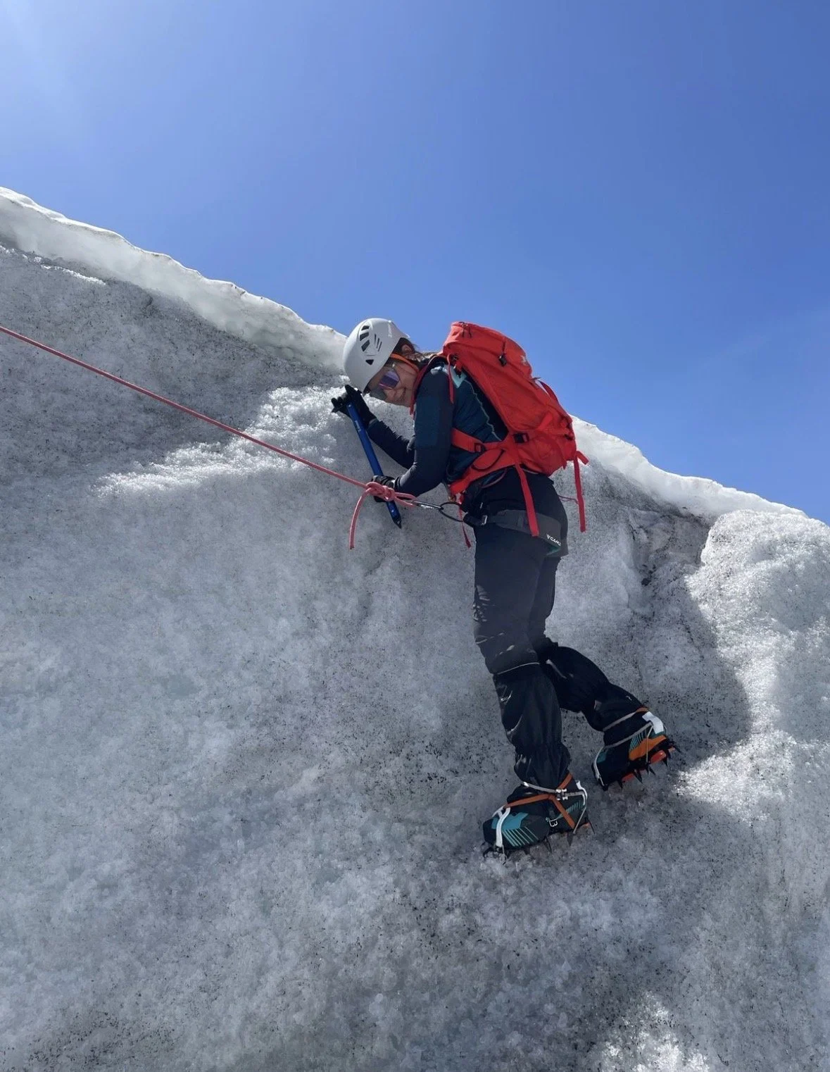 Alpiniste équipé d’un casque, d’un piolet et de crampons escaladant un glacier encordé sous un ciel bleu.