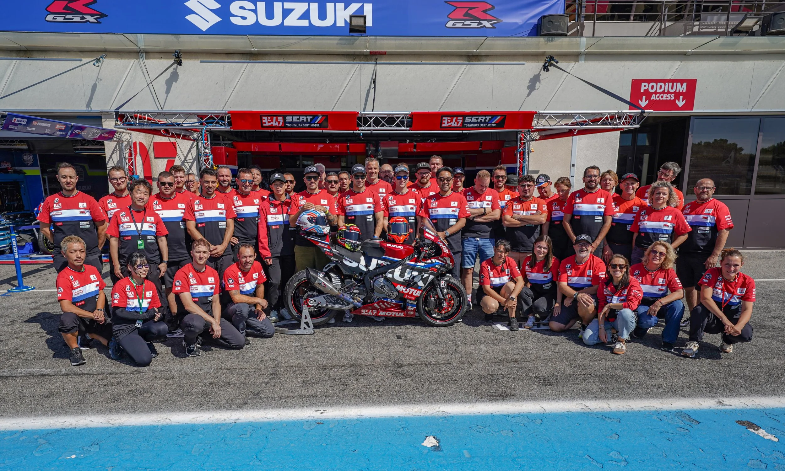 Équipe Yoshimura SERT Motul posant devant une moto Suzuki de course dans un stand de circuit.