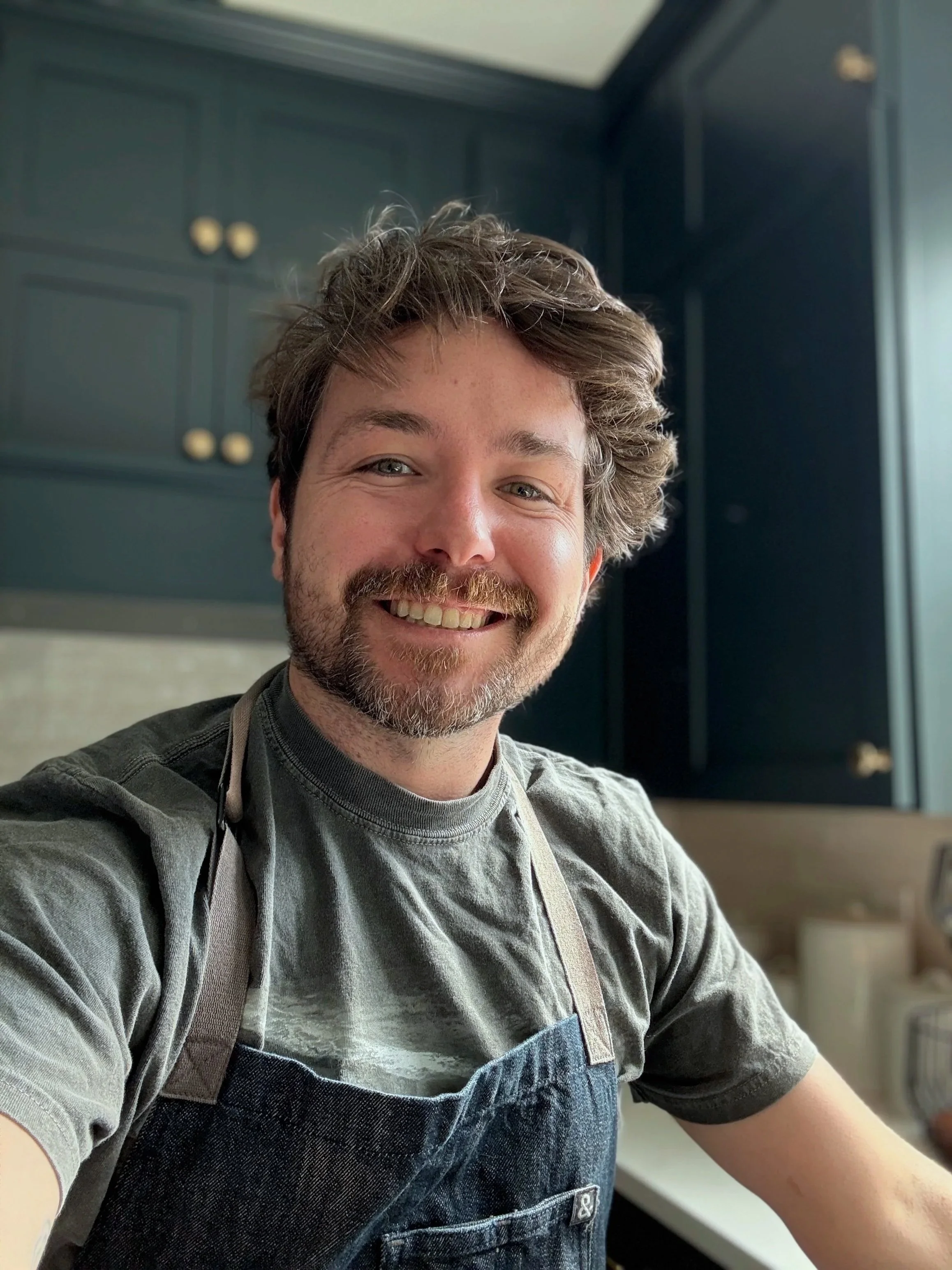 Smiling bearded man wearing a gray shirt and dark denim apron in a bright kitchen