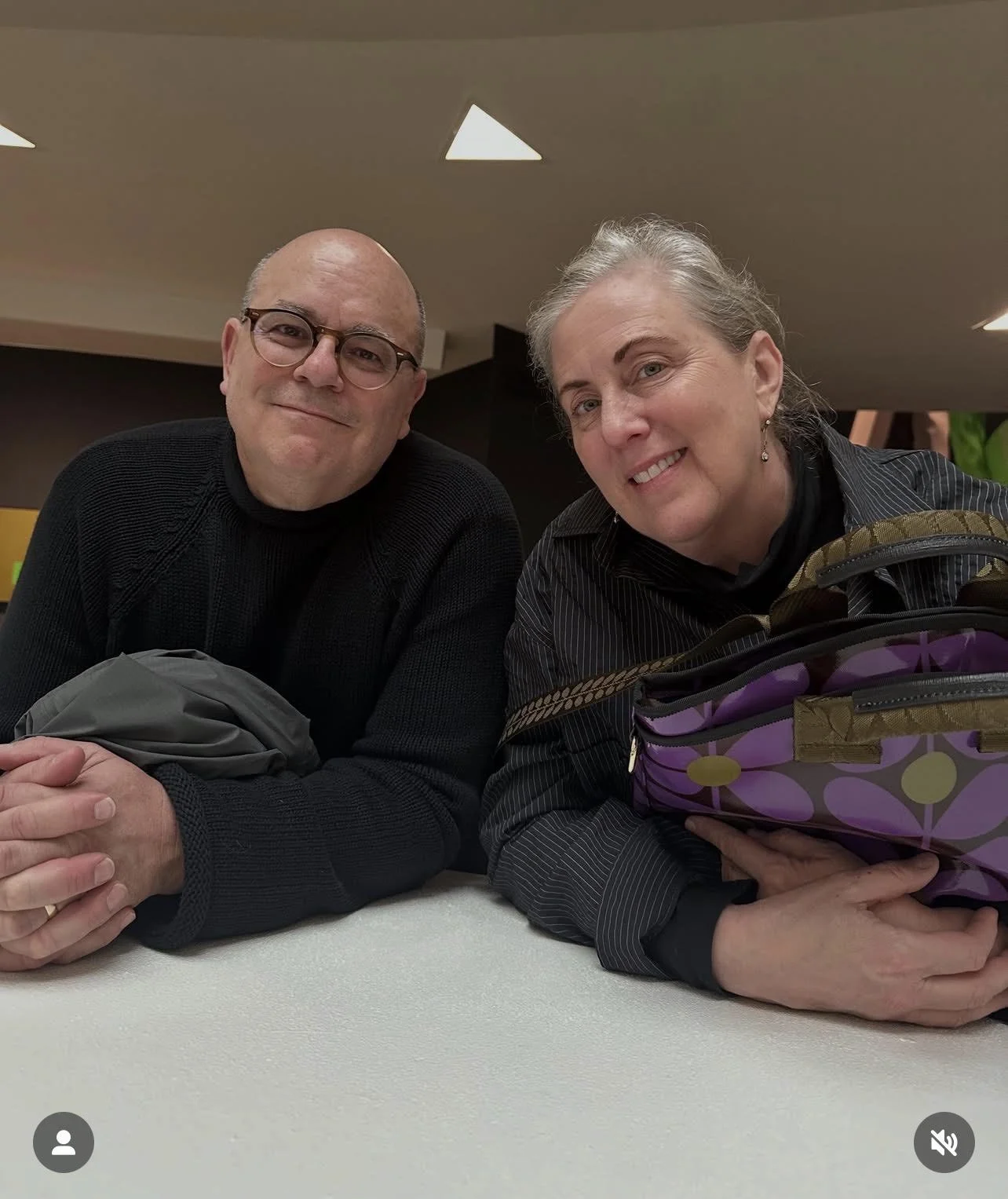 Smiling man and woman seated together at a table, with a colorful patterned bag in the foreground.
