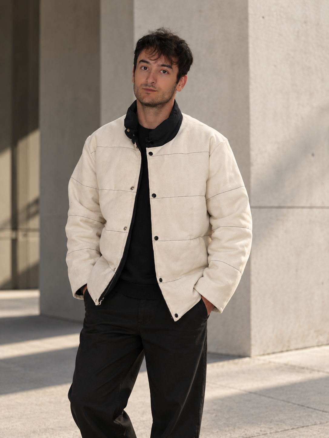 Man wearing a cream jacket and black clothing standing outdoors beside a concrete building