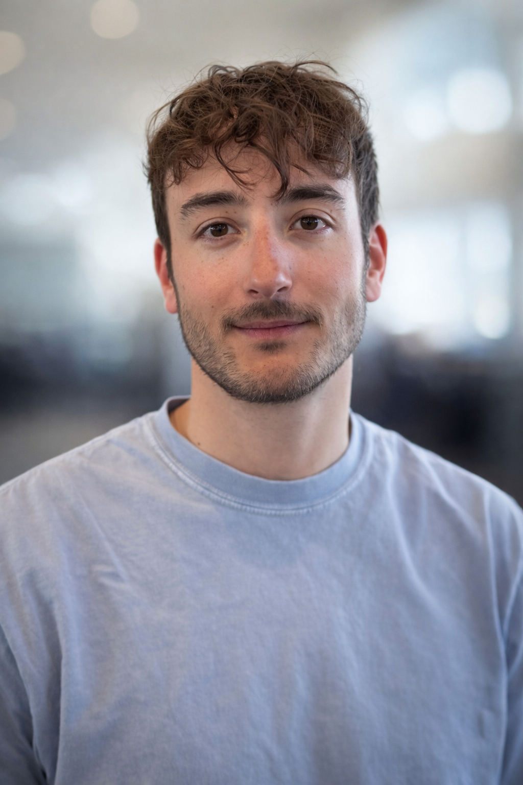 Close-up portrait of a young man with curly brown hair and light beard wearing a pale blue shirt