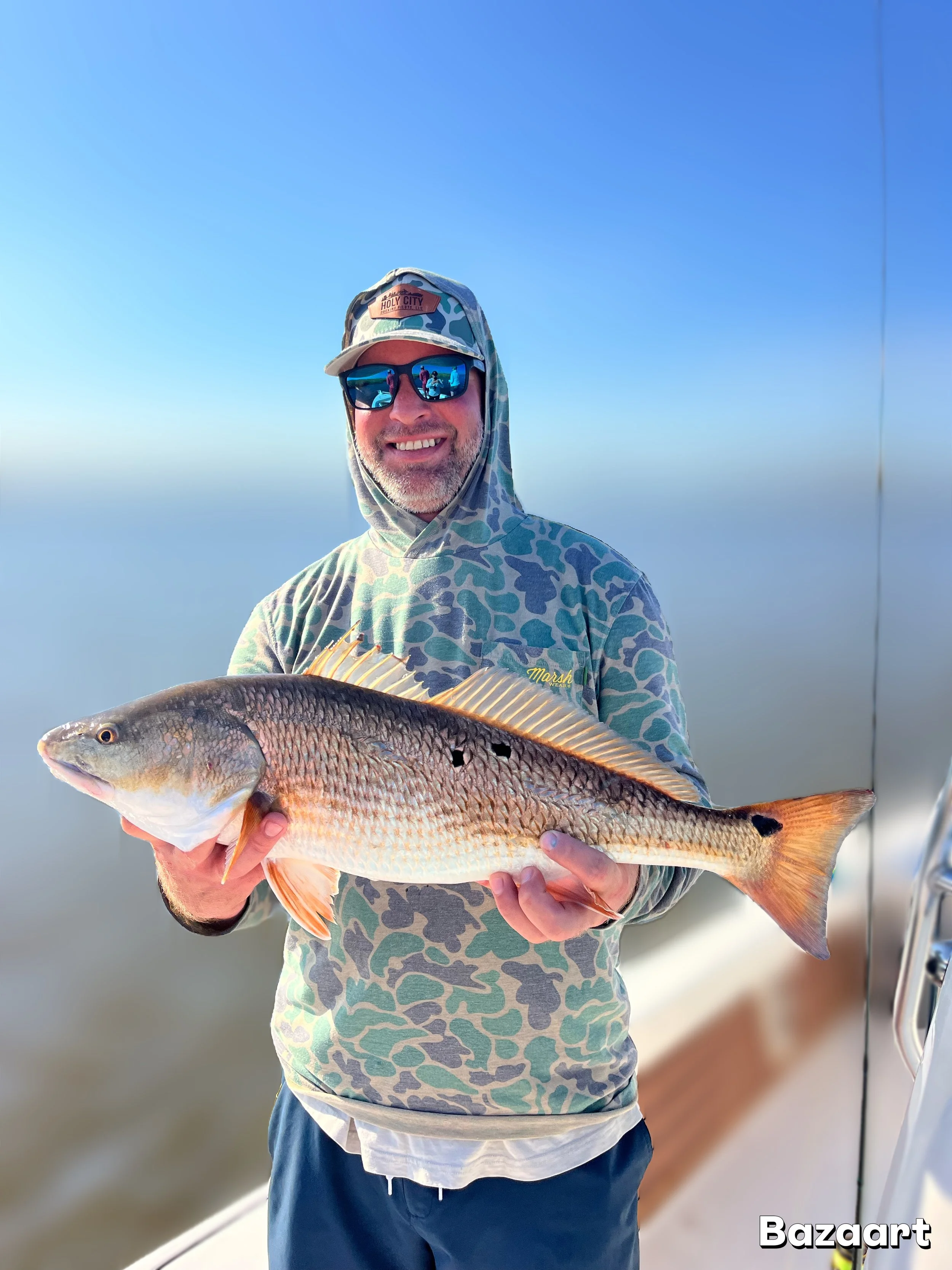 Smiling angler in camouflage clothing and sunglasses holding a large redfish aboard a boat
