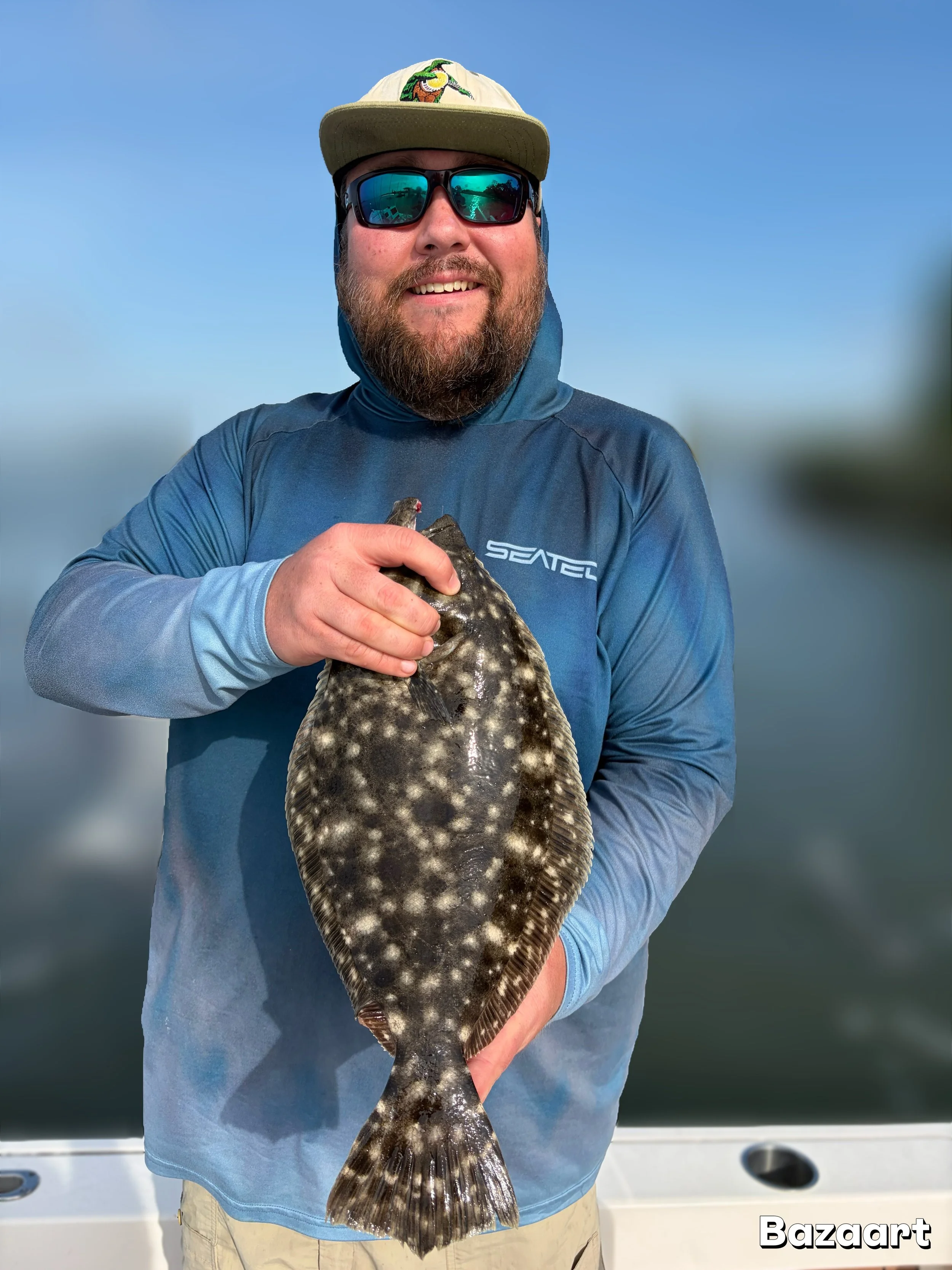 Smiling bearded angler wearing sunglasses and a cap holds a large spotted flatfish on a boat.