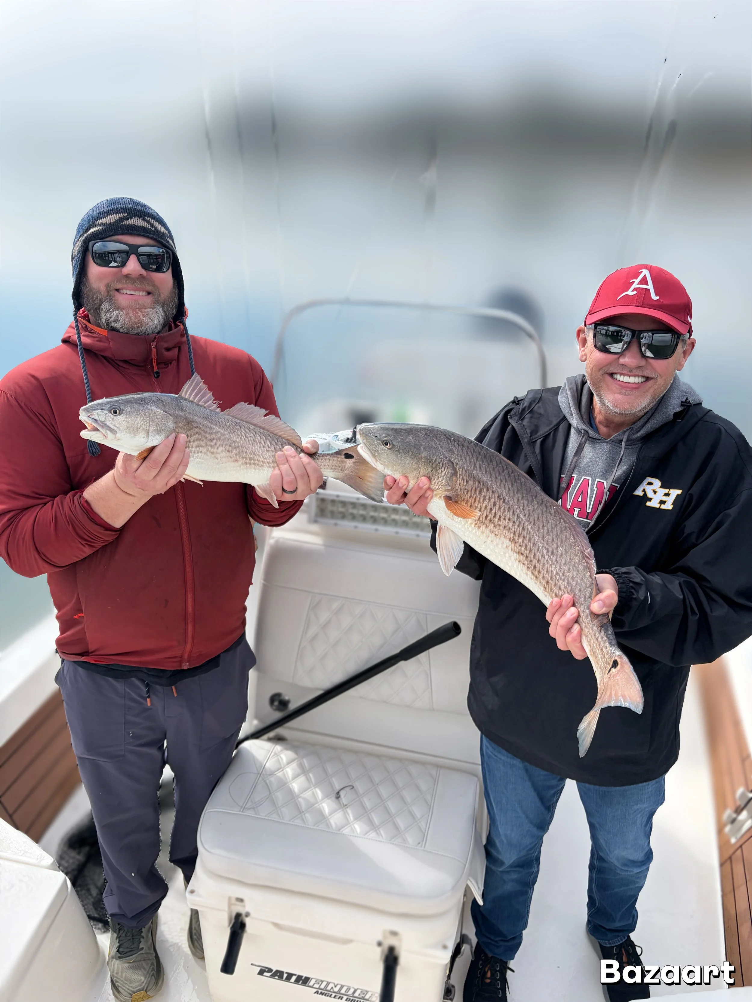 Two anglers wearing sunglasses hold large redfish aboard a fishing boat.