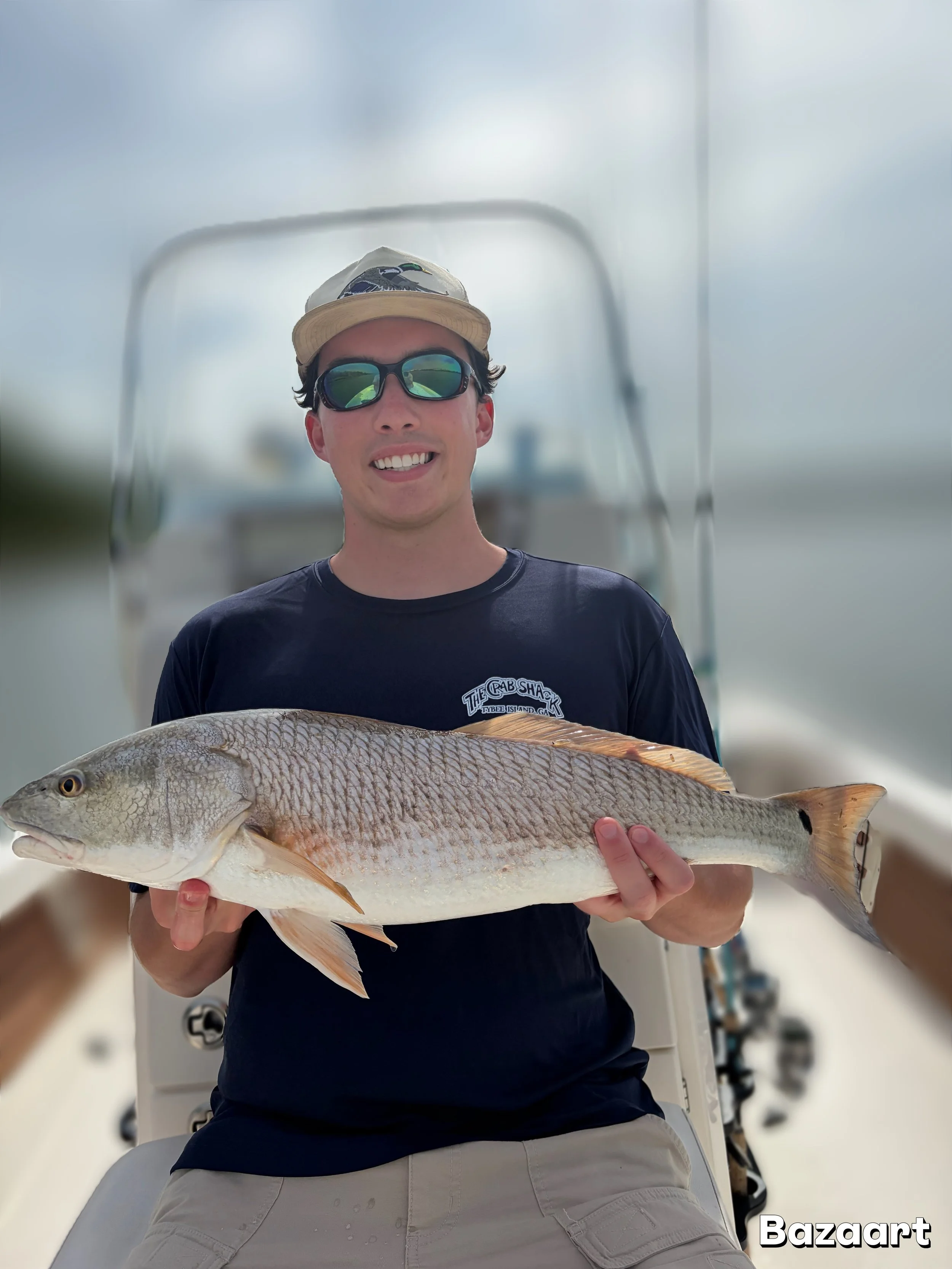 Smiling fisherman wearing sunglasses and a cap holds a large redfish aboard a boat.