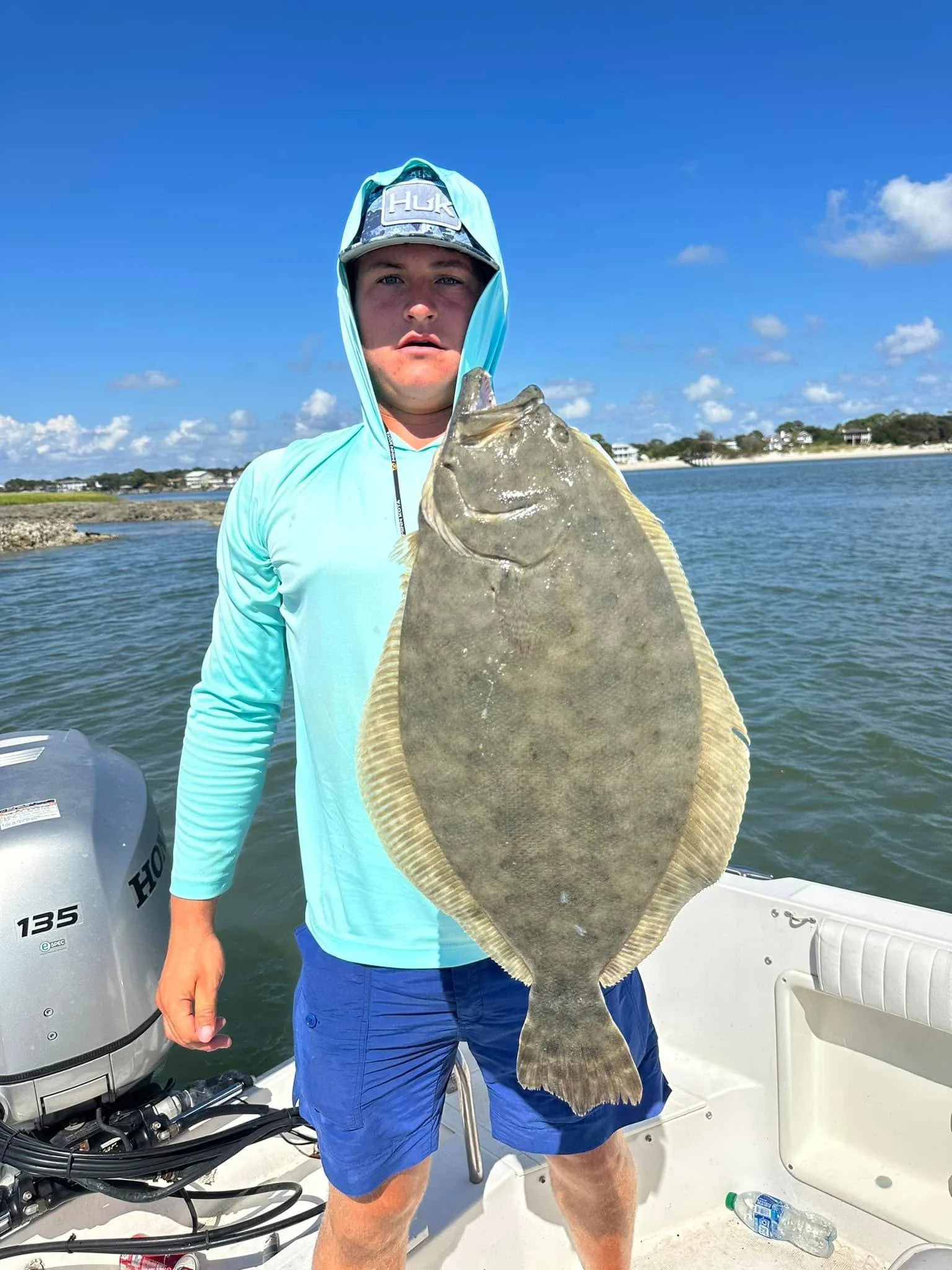 Angler holding a large flounder aboard a fishing boat near a coastal shoreline
