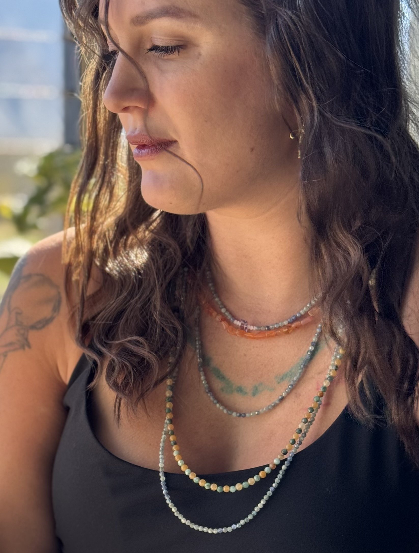 Close-up portrait of a woman with long dark hair, layered beaded necklaces, earrings, and a shoulder tattoo.