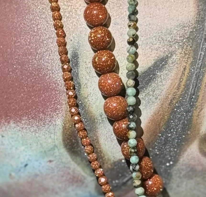 Close-up of three beaded gemstone necklaces, including orange-goldstone beads and multicolored natural stone beads, on a colorful surface.
