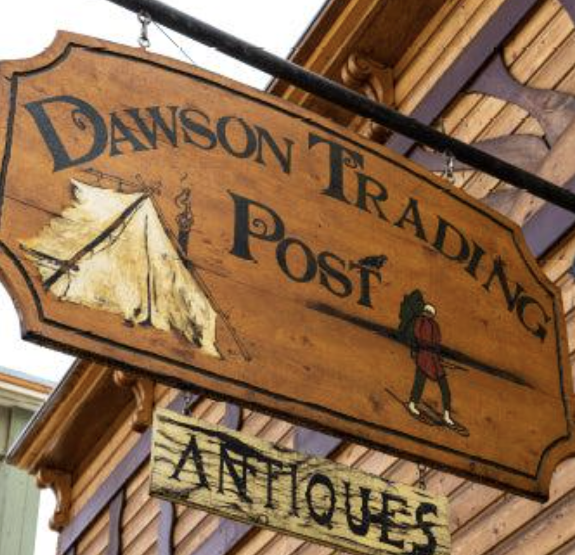 Vintage wooden sign reading “Denniston Trading Post,” illustrated with a tent and hiker, hanging outside a rustic building