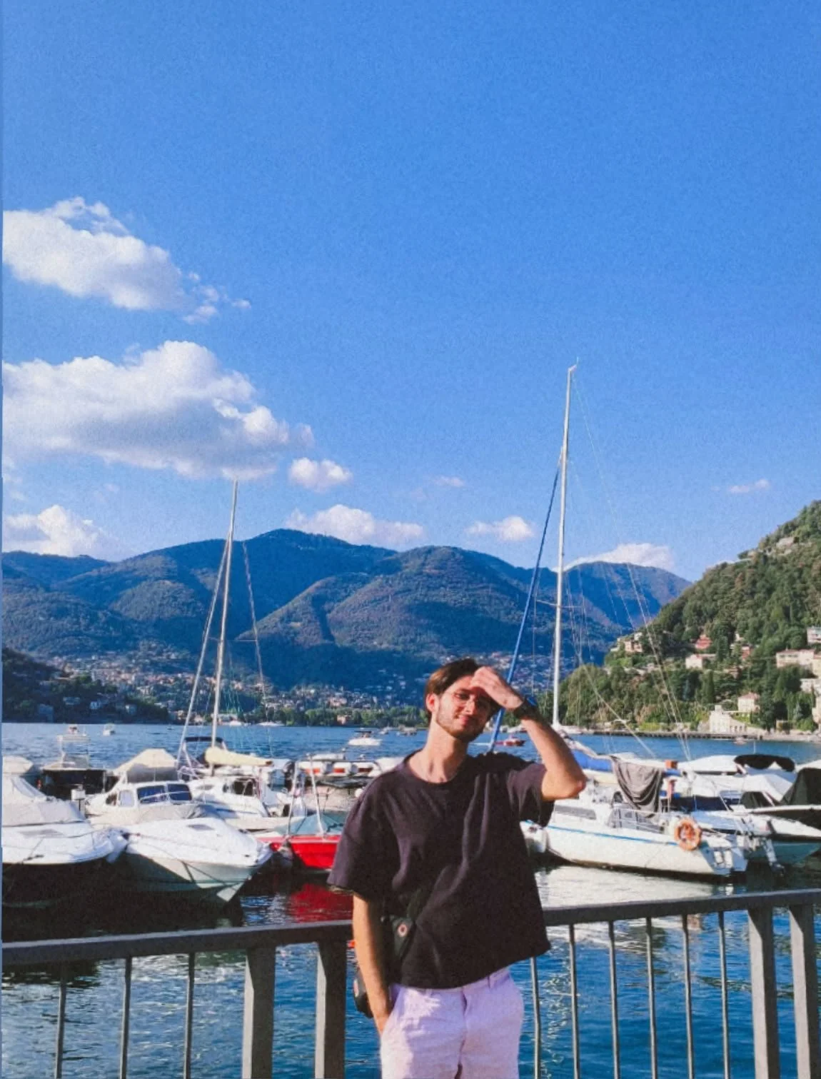 Man standing by a marina with sailboats, mountains, and blue sky in the background