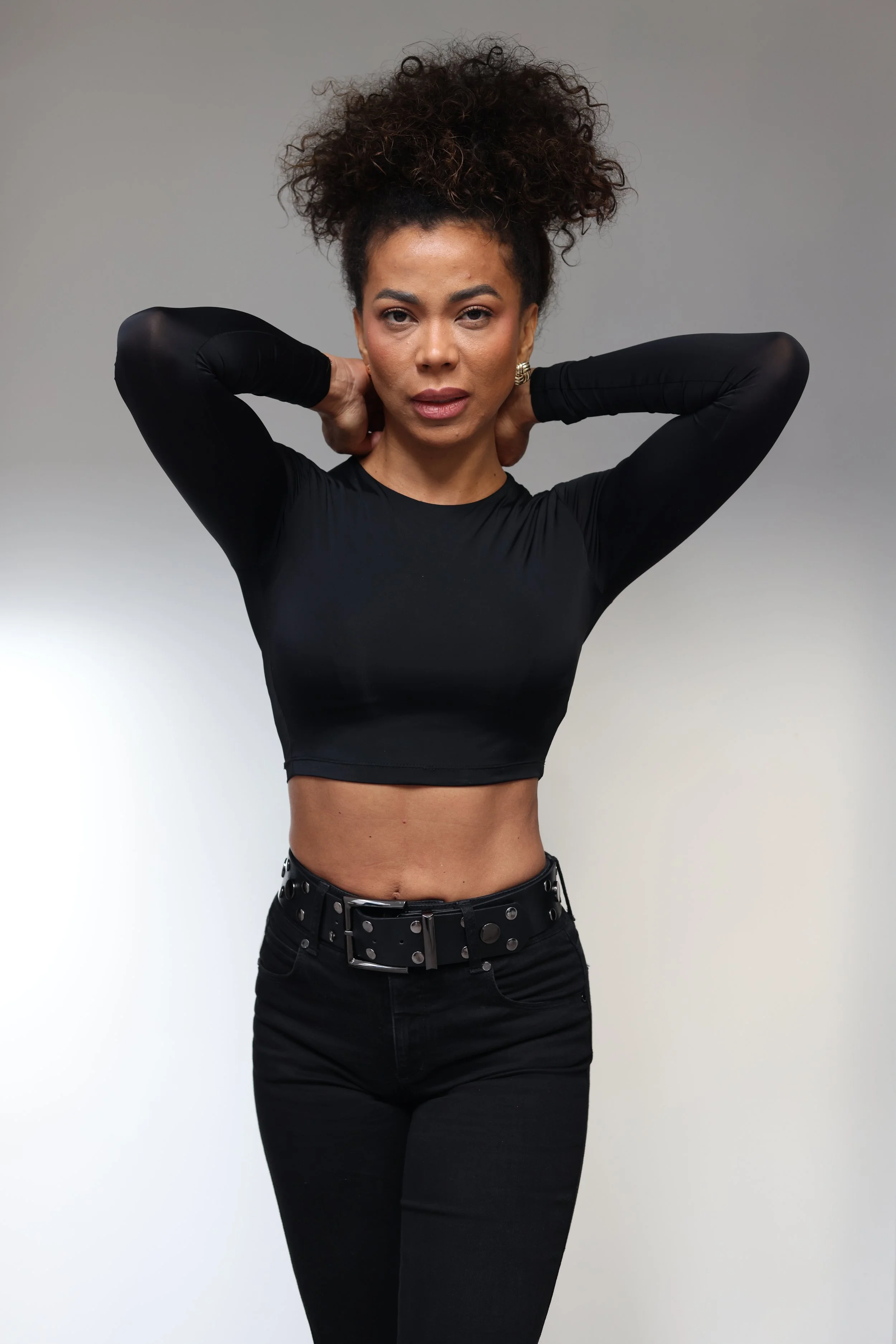 Woman with curly hair wearing a black top and jeans, posing with her hands behind her neck against a gray background