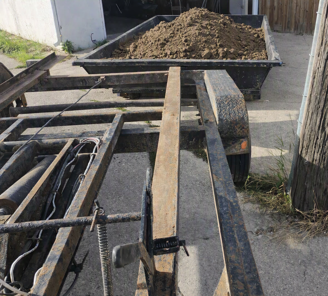 Rusty utility trailer loaded with soil outside a garage