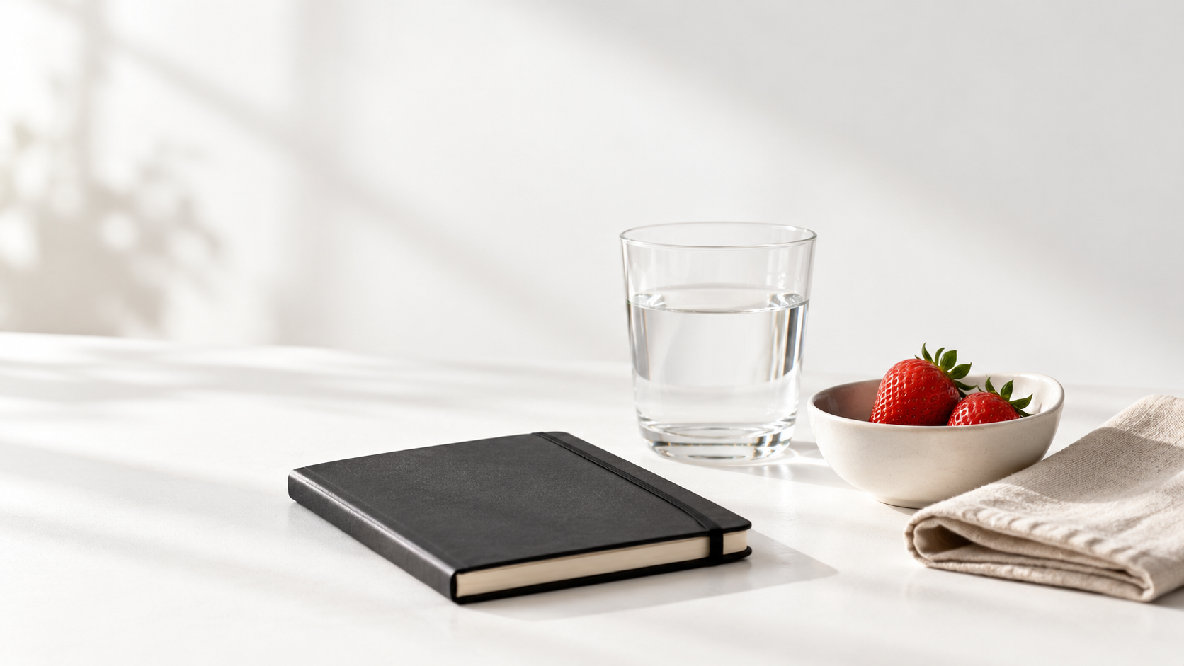 AI placeholder: glass of water, strawberries, a black notebook, and folded linen on a bright white table.