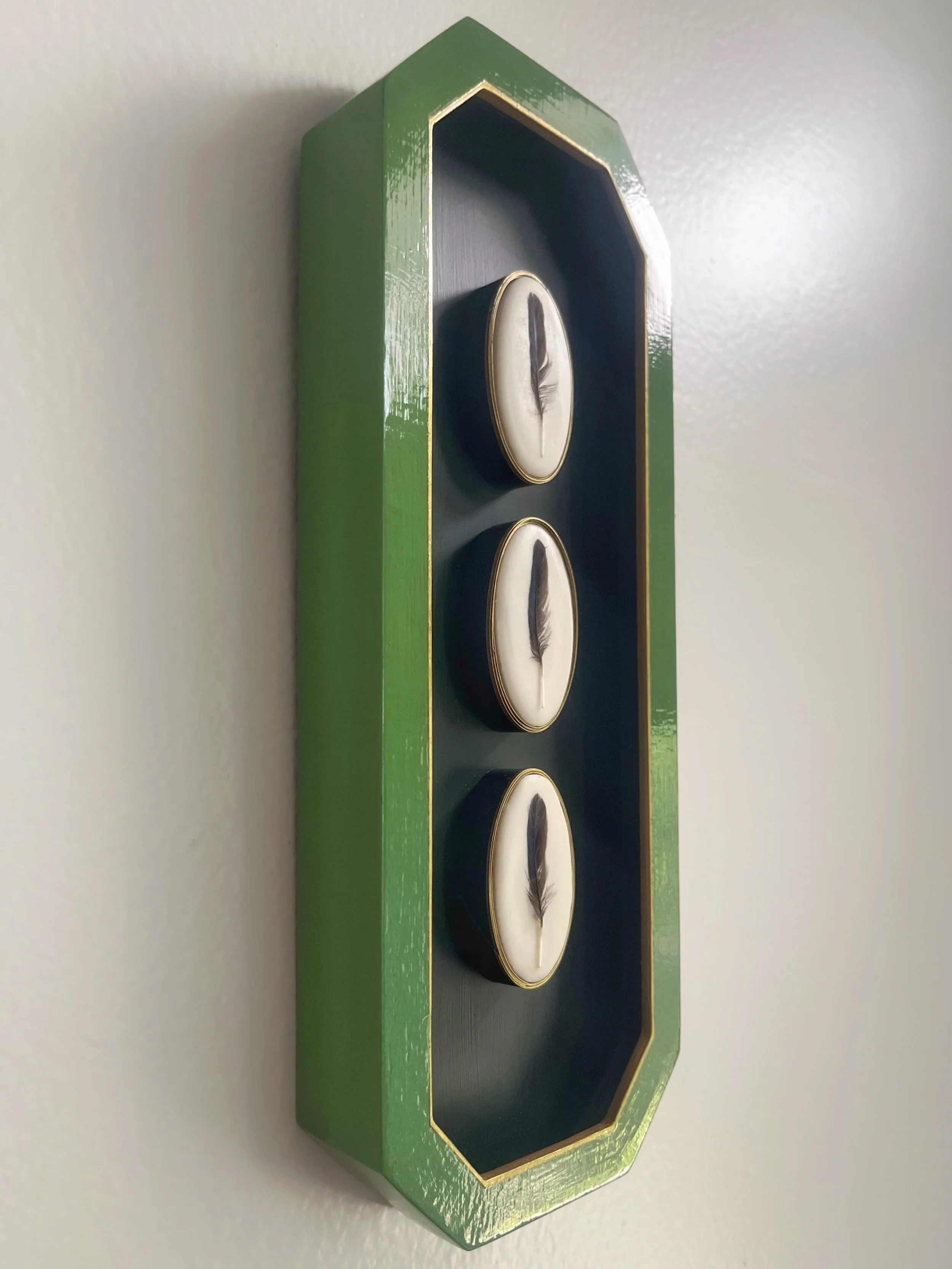 Green, gold-trimmed wall-mounted shadow box displaying three white feathers on a dark blue background