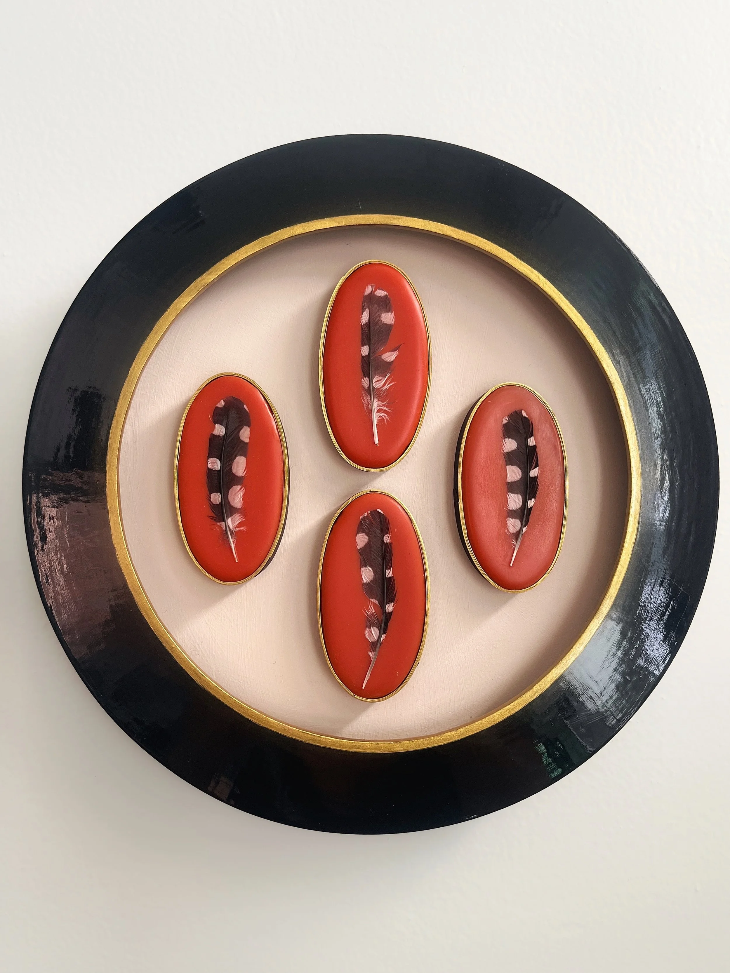 Round black and gold decorative wall plaque featuring four red oval panels with feather motifs