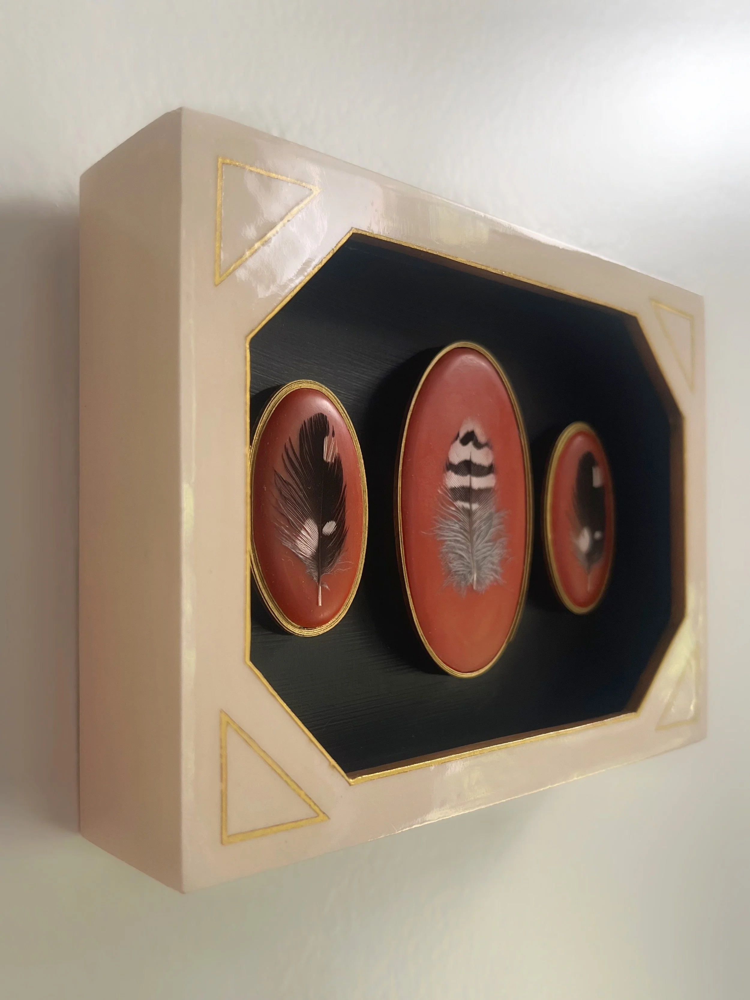 Framed display of three decorative feather paintings on red backgrounds with gold trim.