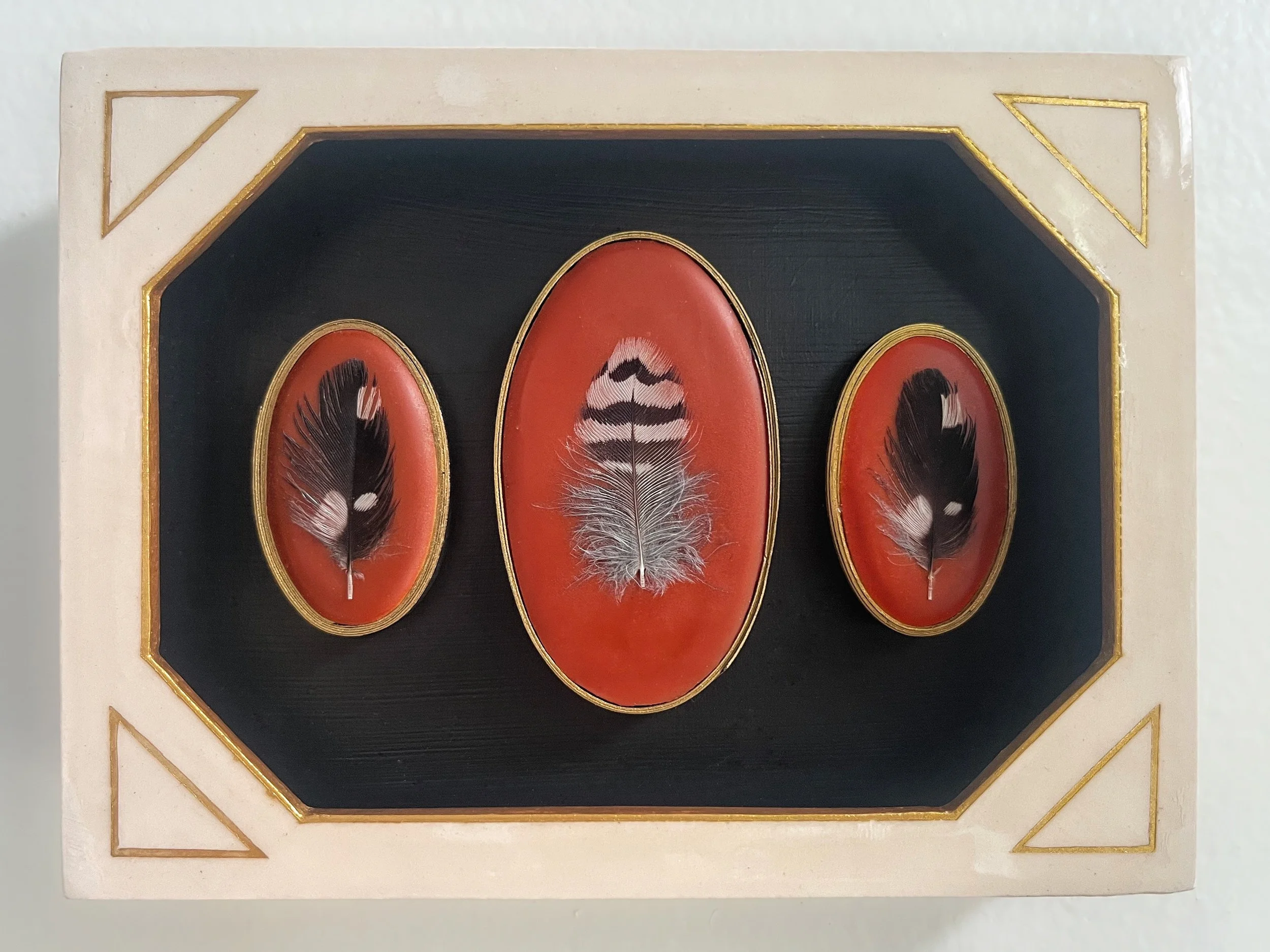 Framed decorative artwork displaying three black-and-white feathers on red oval panels with gold trim.
