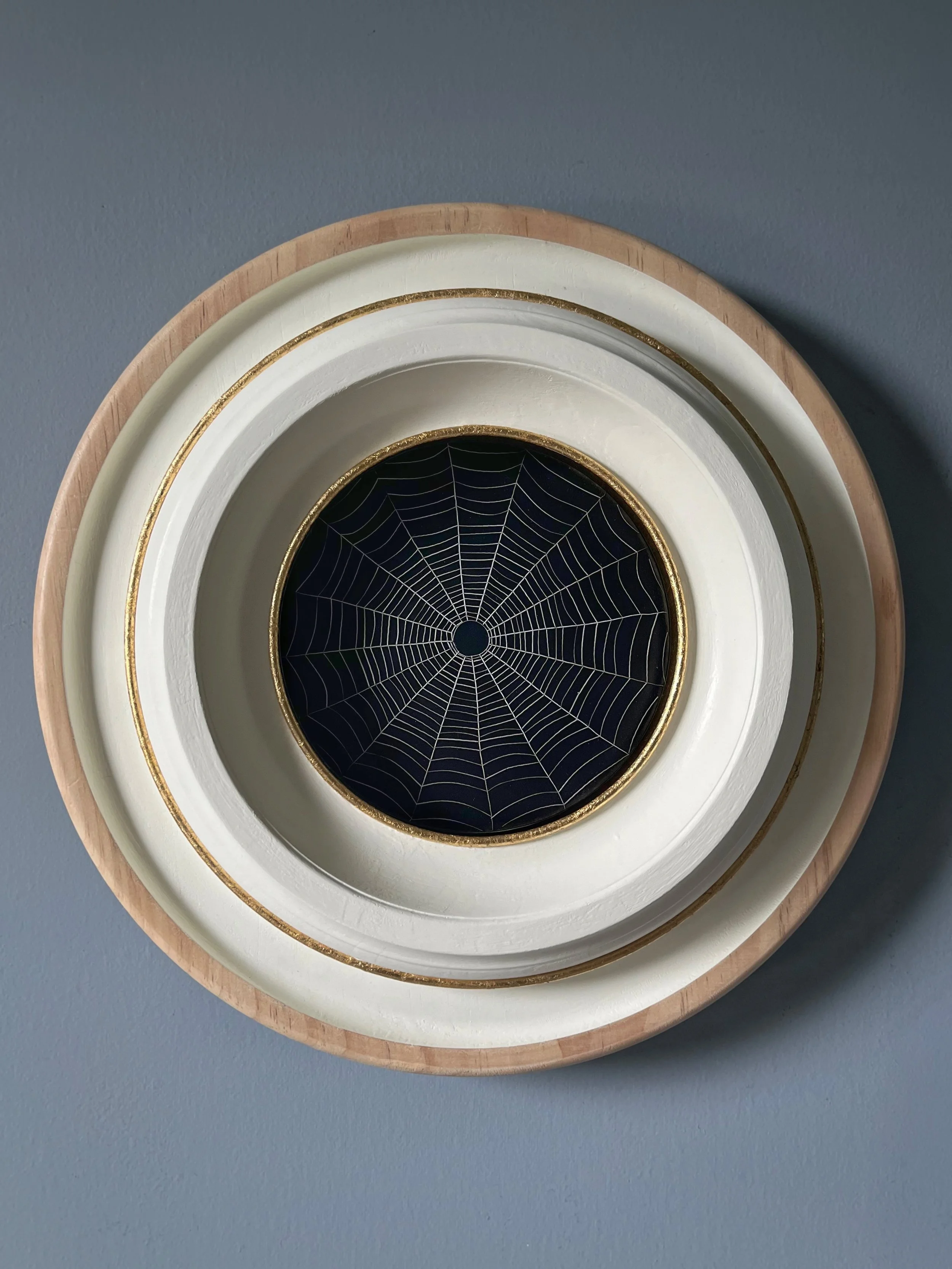 Close-up of a white and gold decorative circular wall plate with a black spiderweb design, set in a wooden frame against a blue wall.