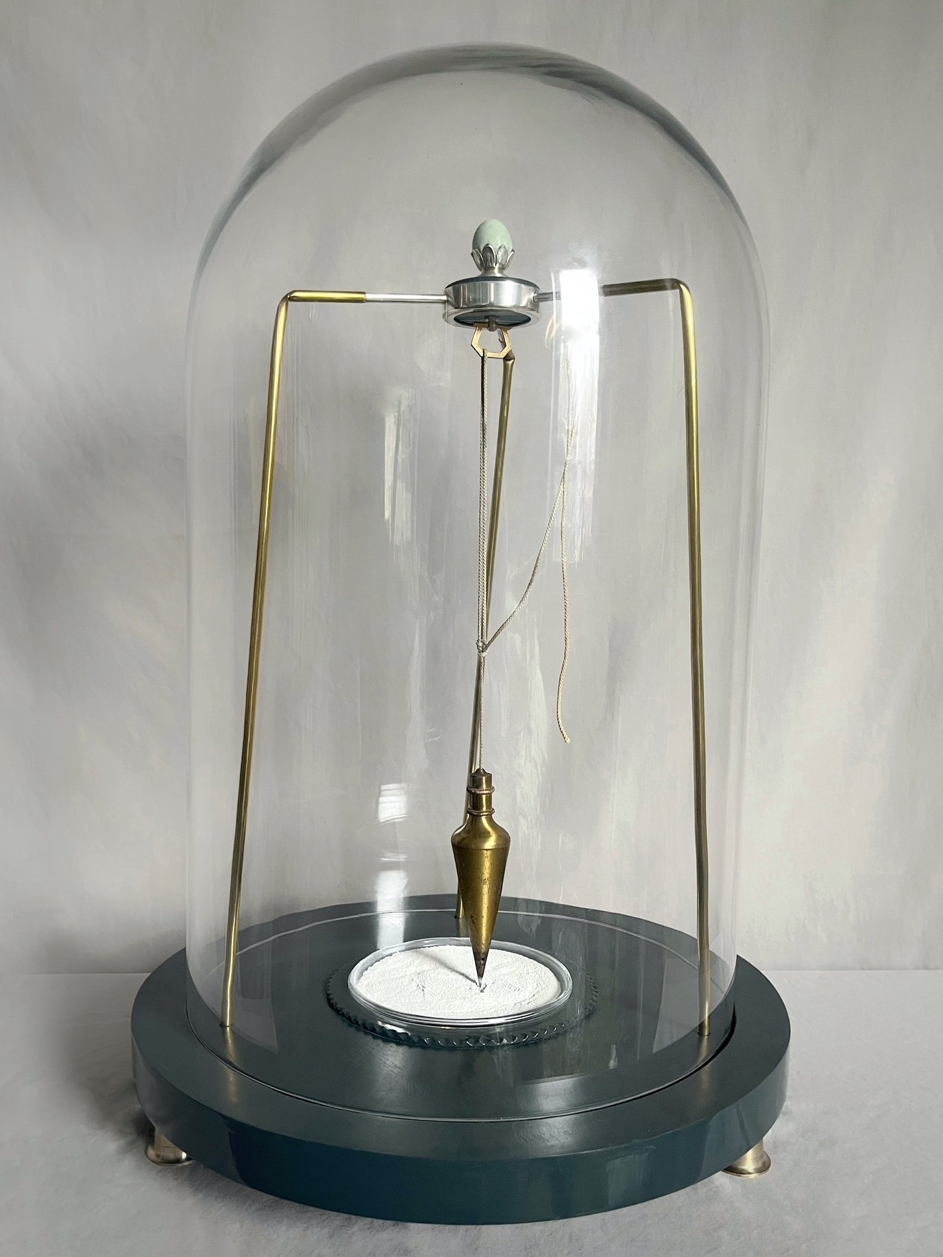 Foucault pendulum with a brass bob suspended over a circular sand tray inside a glass dome