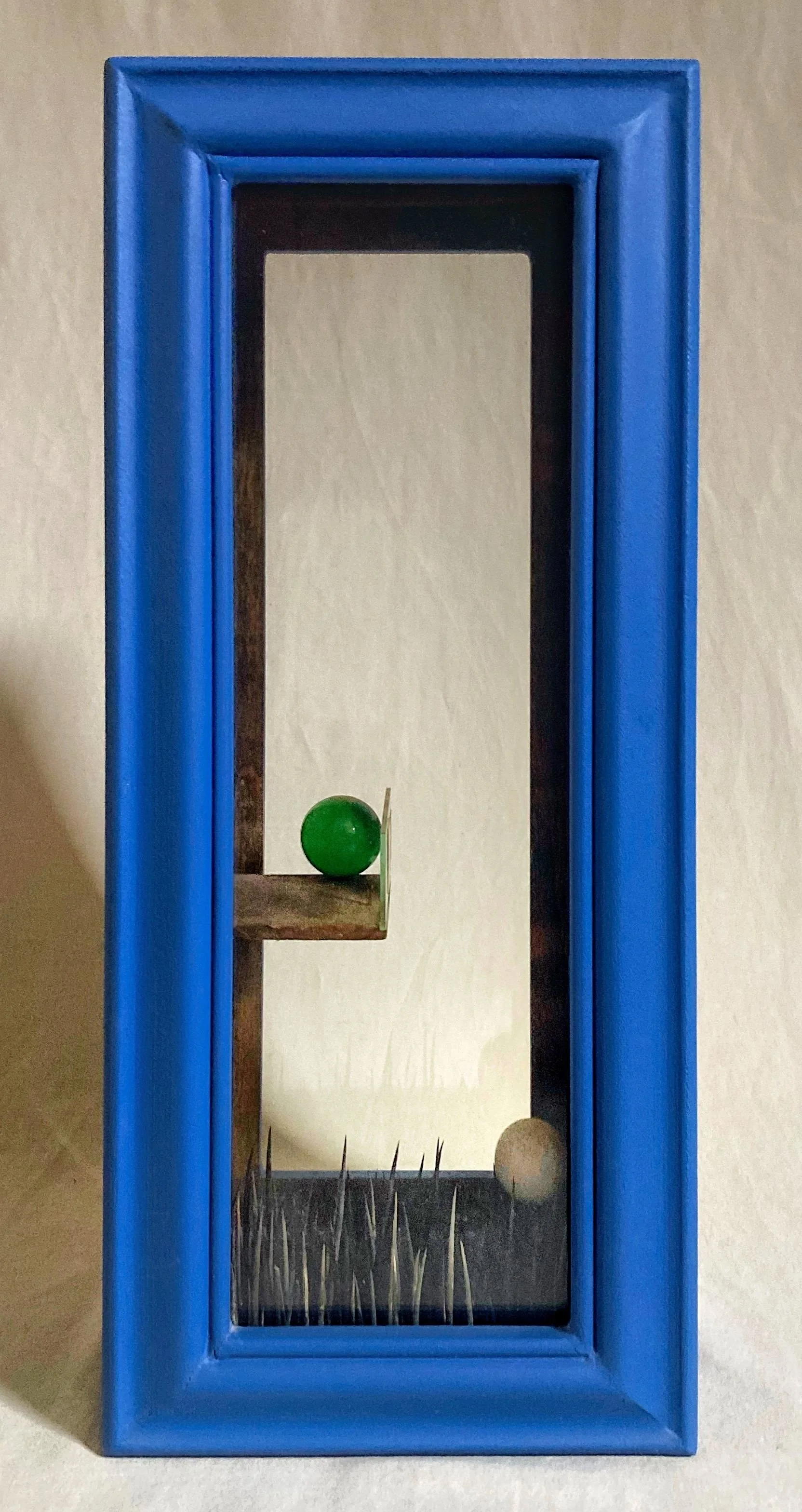Blue framed artwork depicting a green ball, beige ball, and tall grass-like shapes on abstract geometric panels