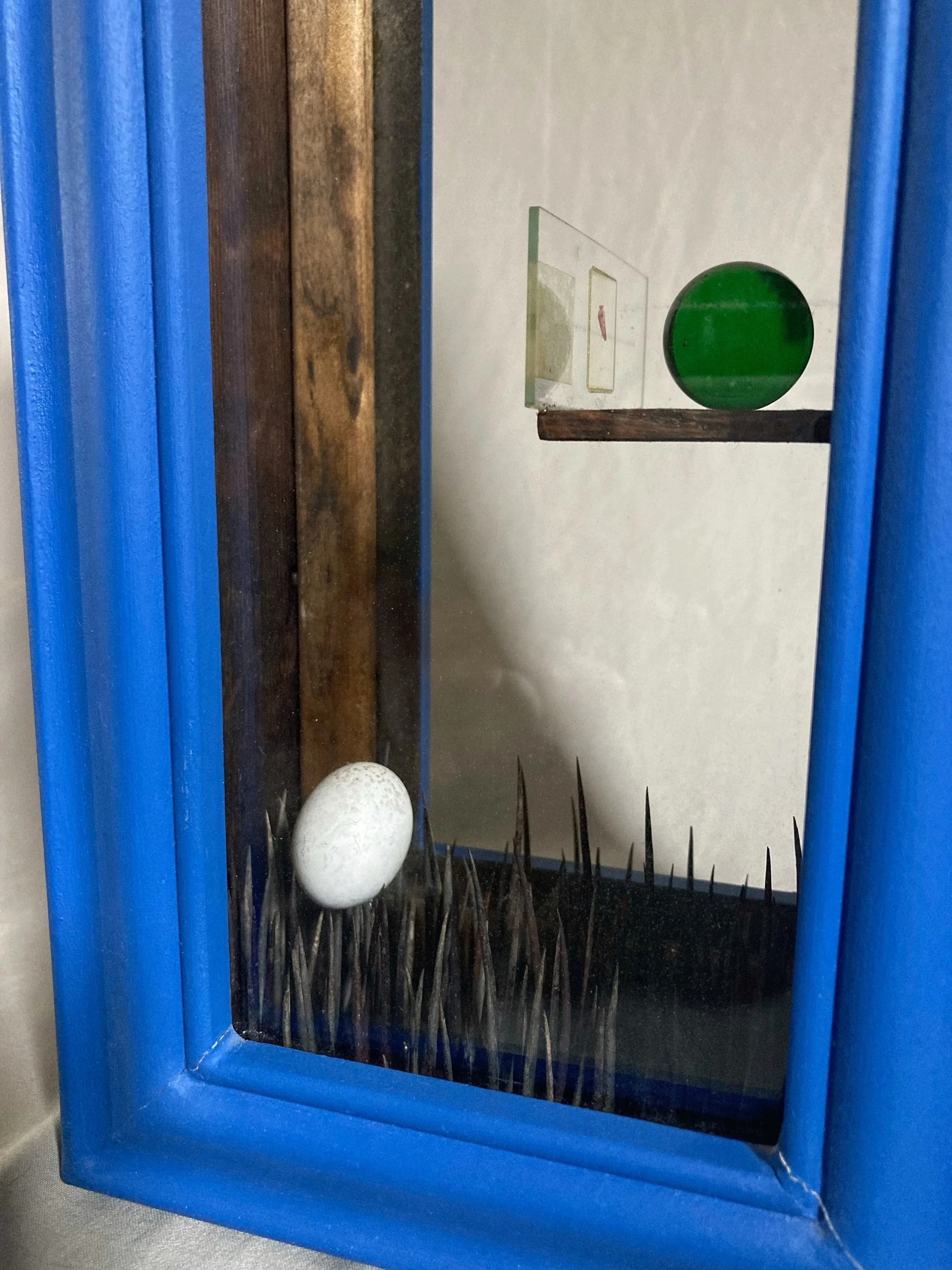 Abstract framed artwork with a green sphere and white golf ball on a black spiked surface, surrounded by a bright blue frame.