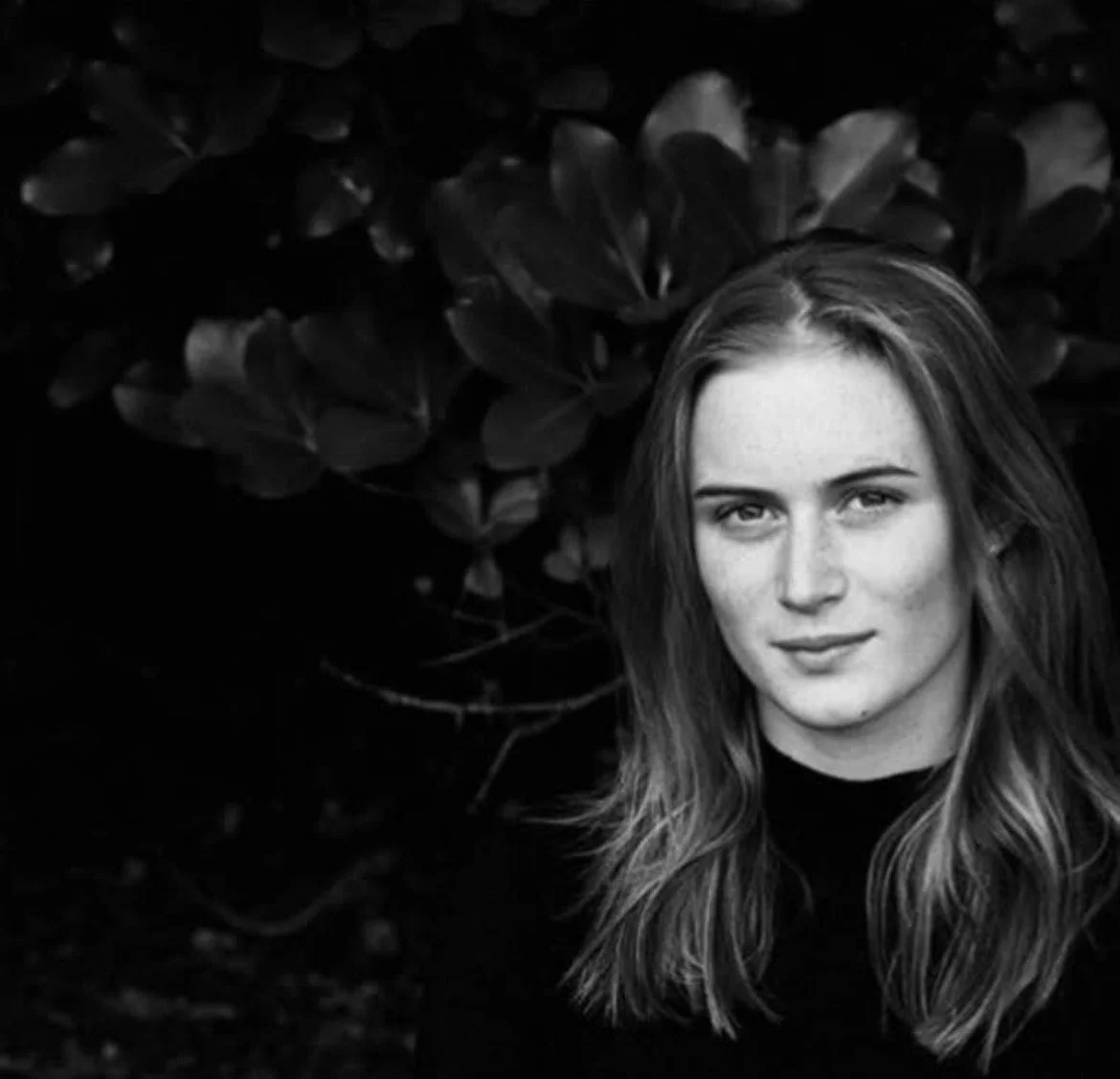 Black-and-white portrait of a woman with long hair standing in front of leafy foliage