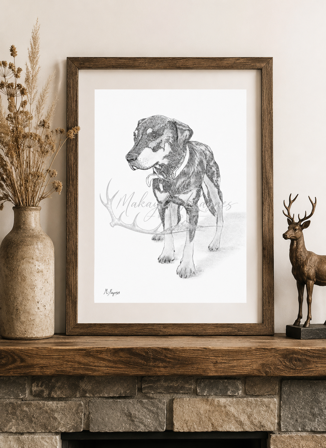 Framed pencil drawing of a dog displayed on a rustic stone fireplace mantel beside dried flowers and a bronze deer statue