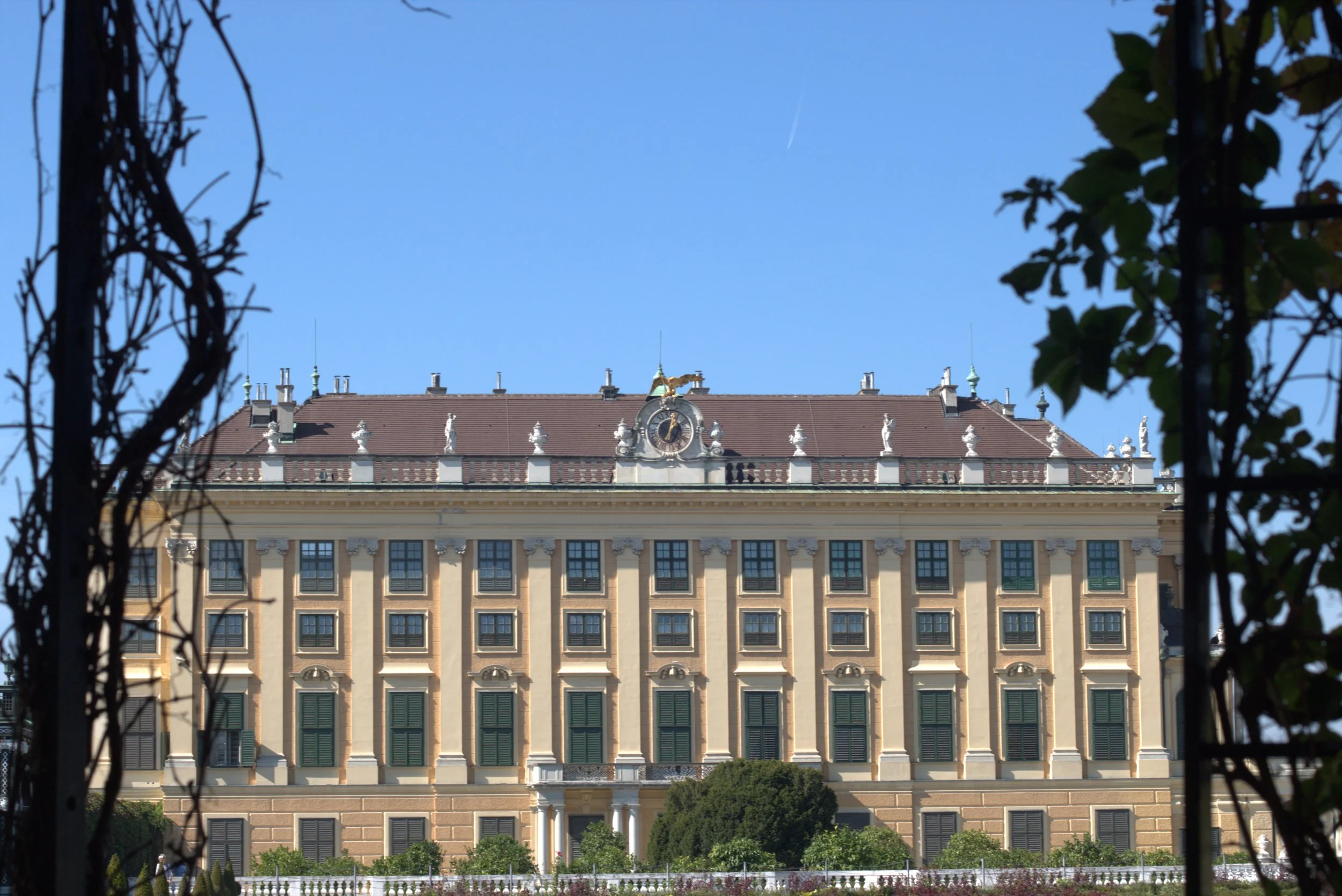 Ornate historic palace facade with a central clock and rooftop golden eagle, framed by foliage under a clear blue sky
