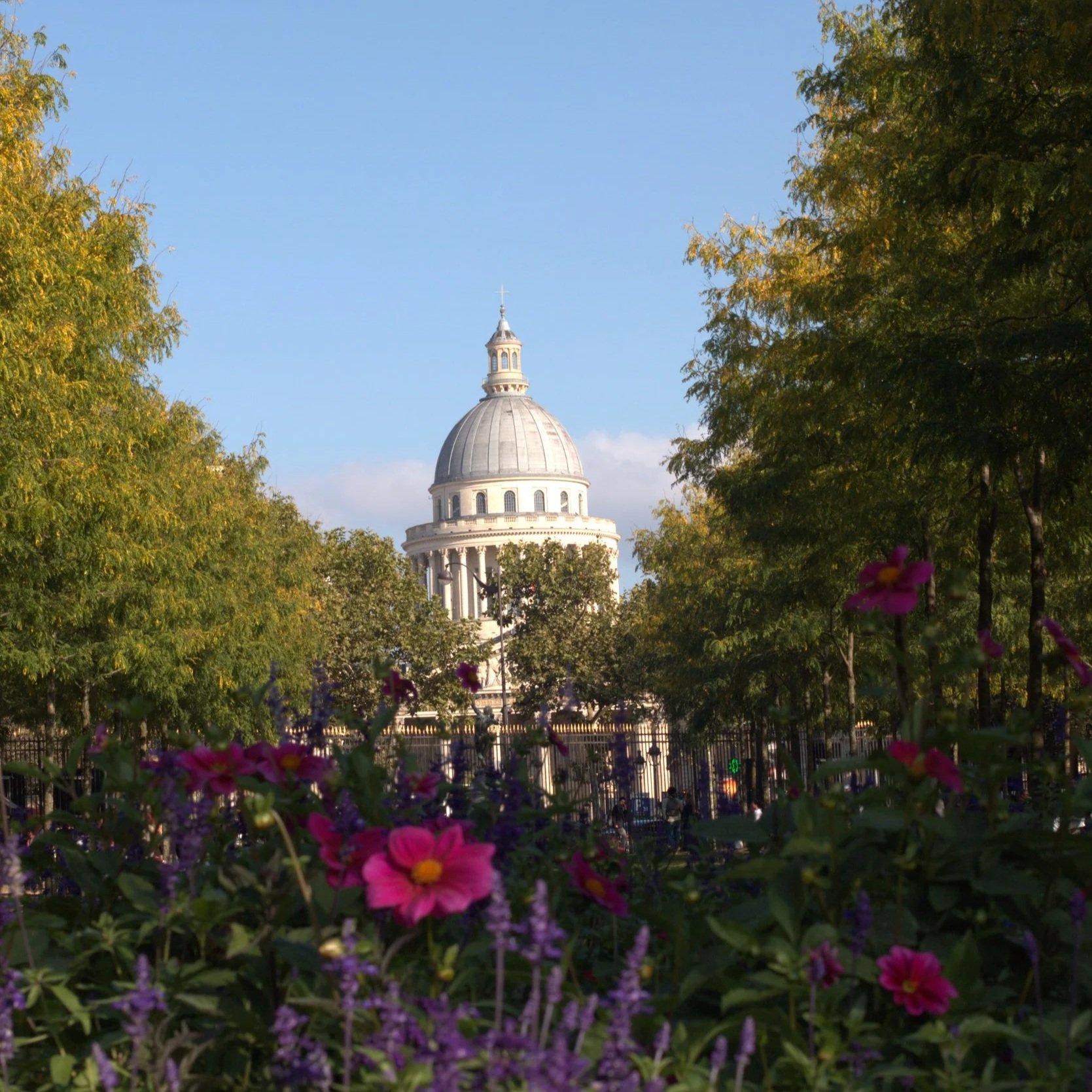Domed historic building with a cross, framed by trees and colorful garden flowers