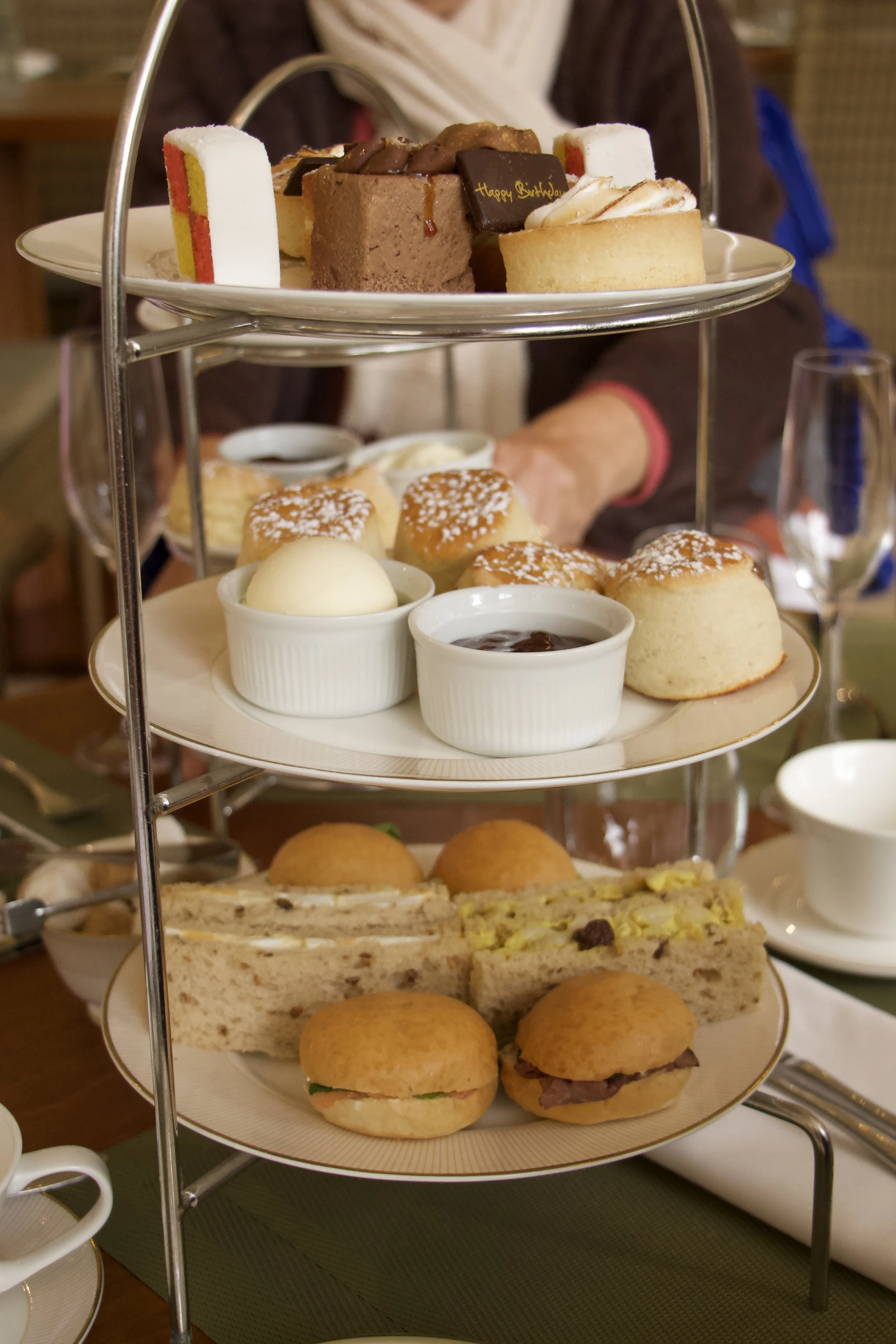 Three-tier afternoon tea stand with cake, pastries, sandwiches, and scones with cream, and jam.