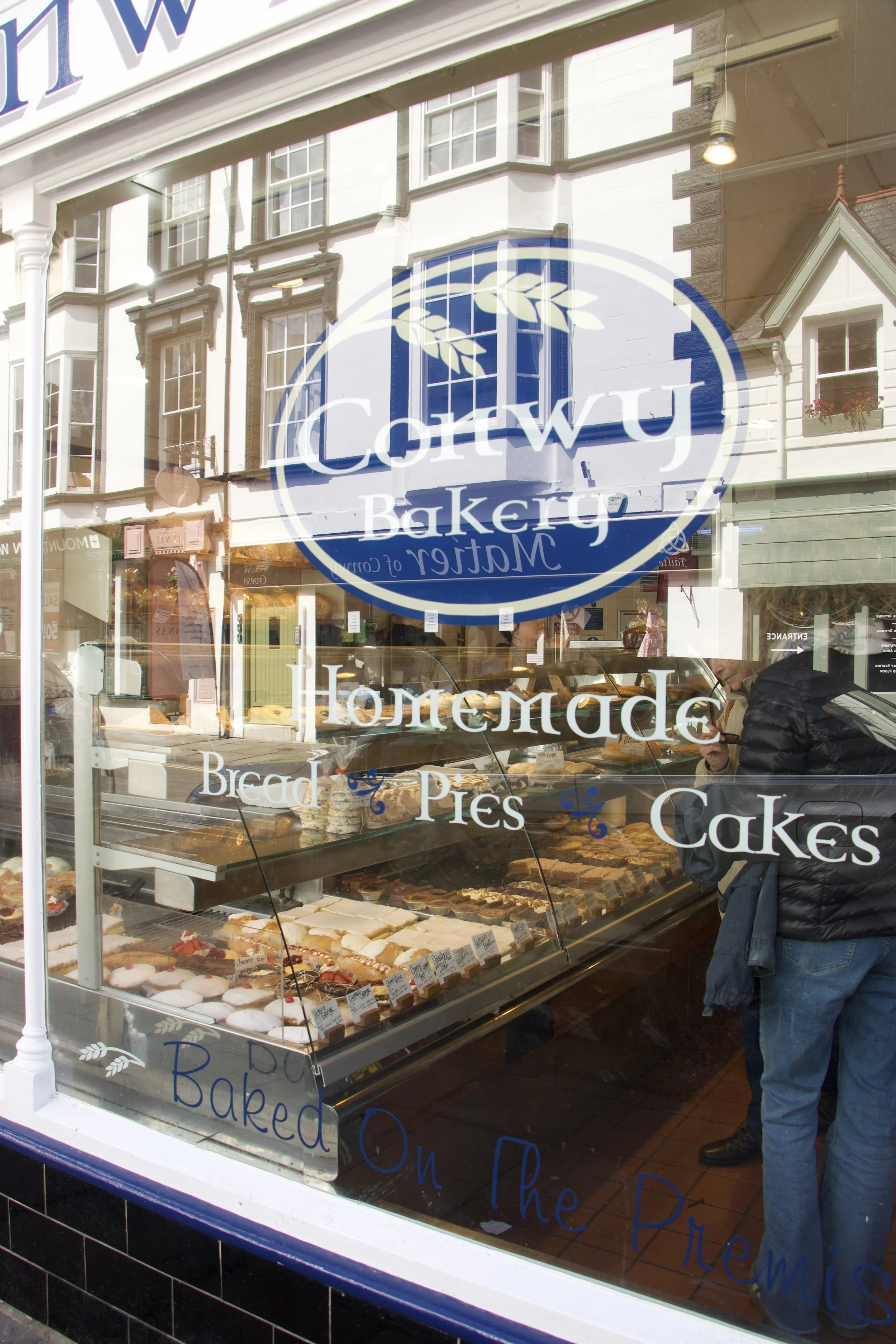 Conwy bakery storefront window displaying cakes, pastries, bread, and a large blue-and-white bakery logo.