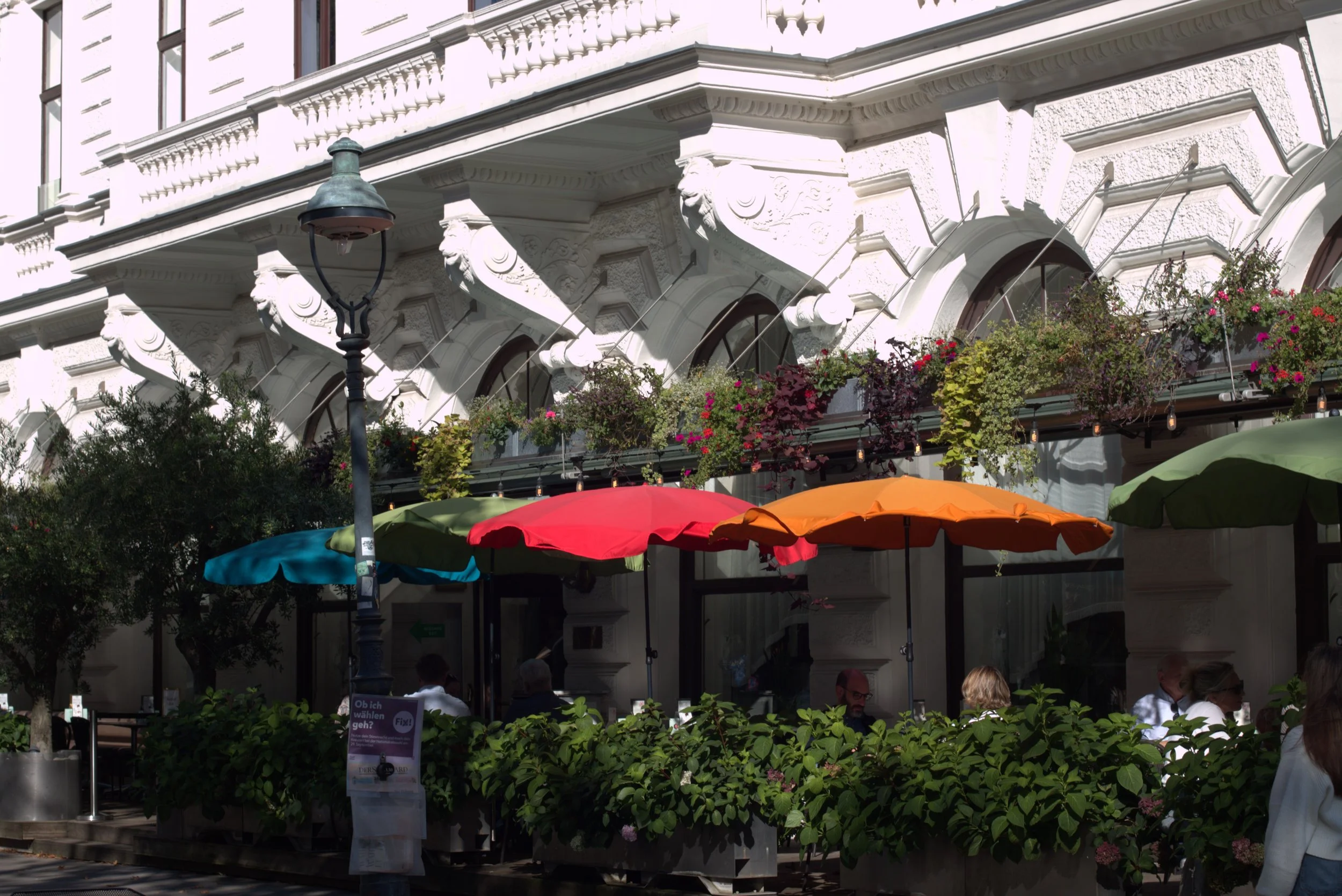 Outdoor restaurant terrace with colorful umbrellas, hanging flower baskets, and an ornate white historic building facade