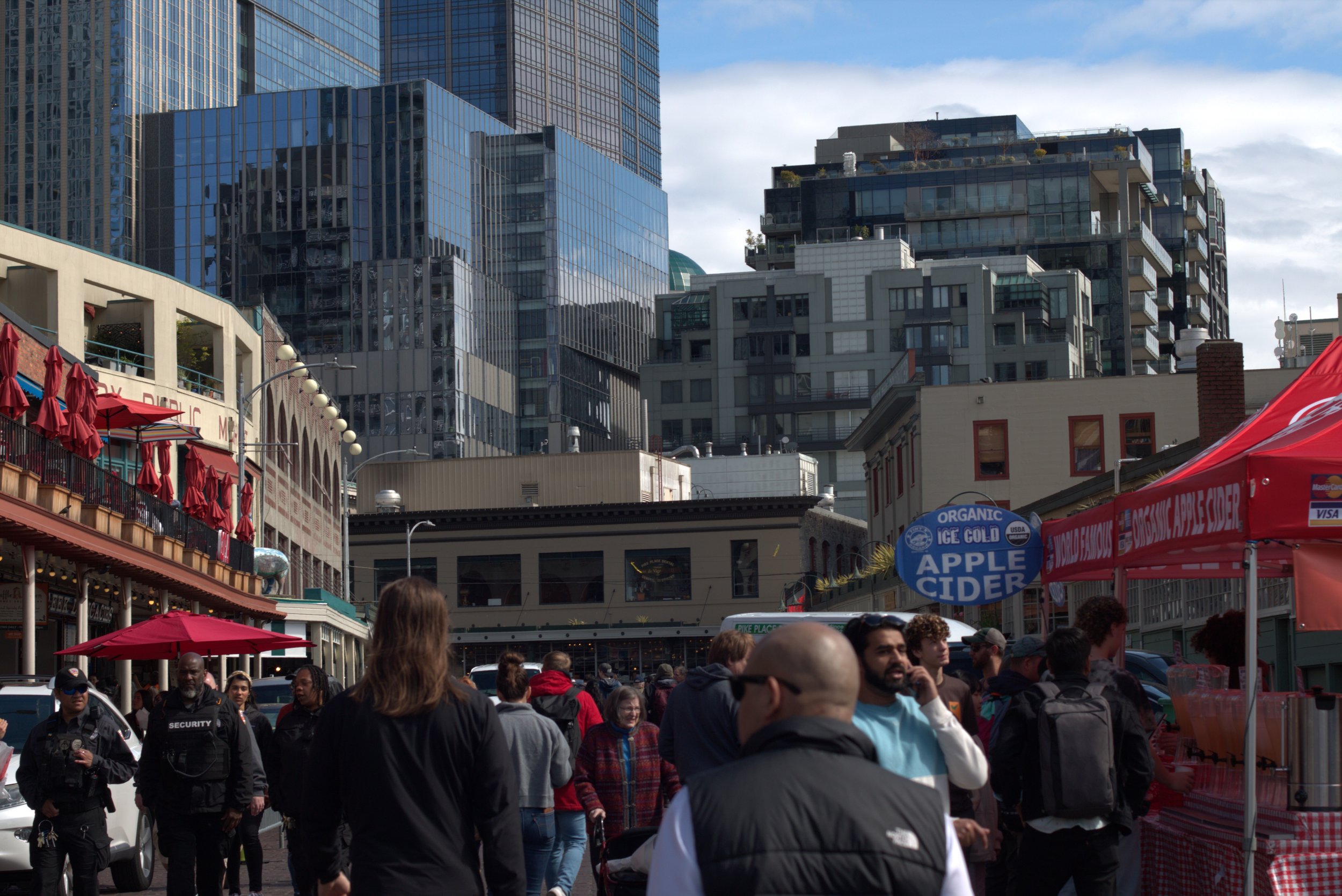 Crowded Pike Place Market in Seattle with red umbrellas, food vendors, shoppers, and downtown high-rise buildings
