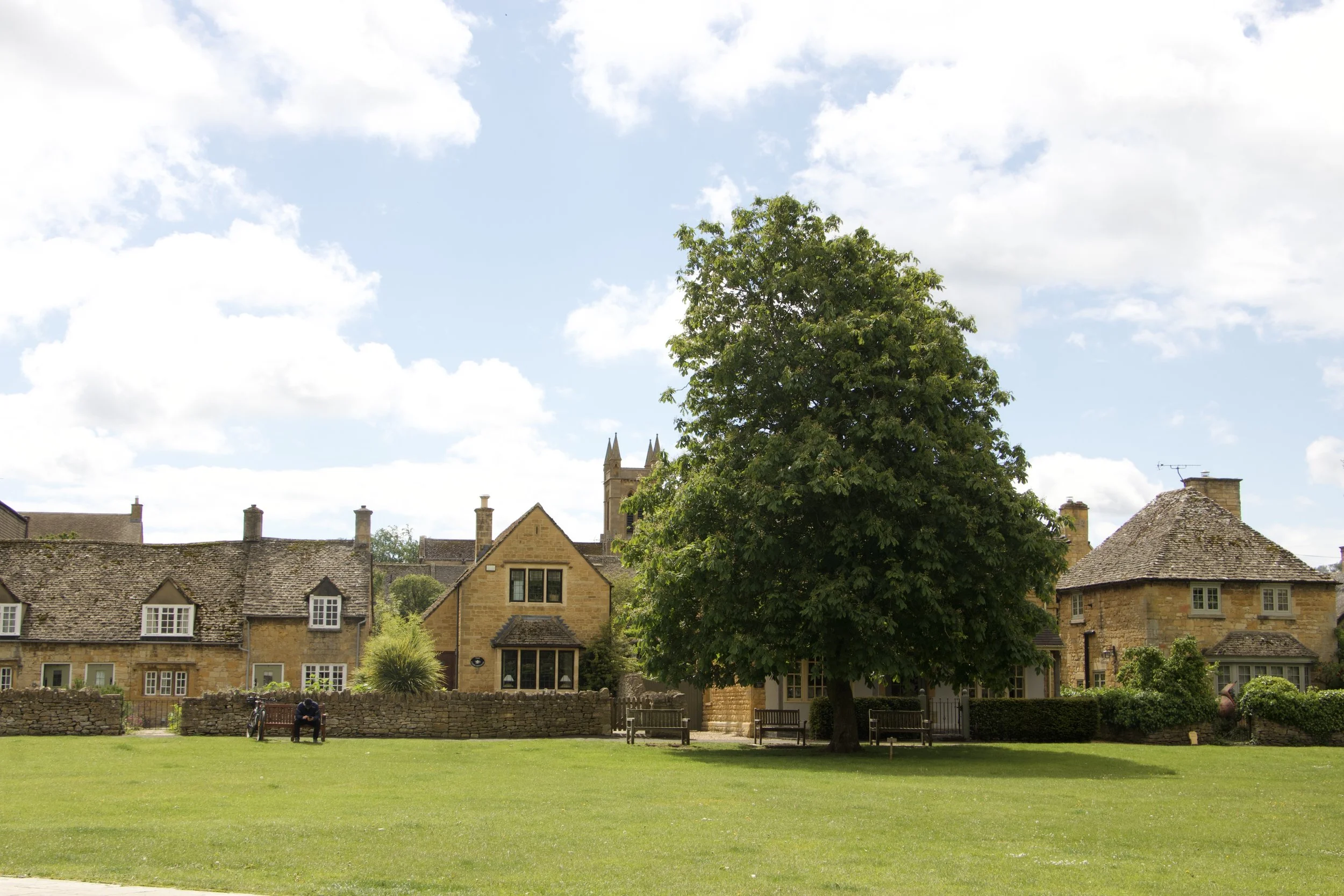 Traditional honey-colored stone cottages with slate roofs beside a village green and large leafy tree under a partly cloudy blue sky.