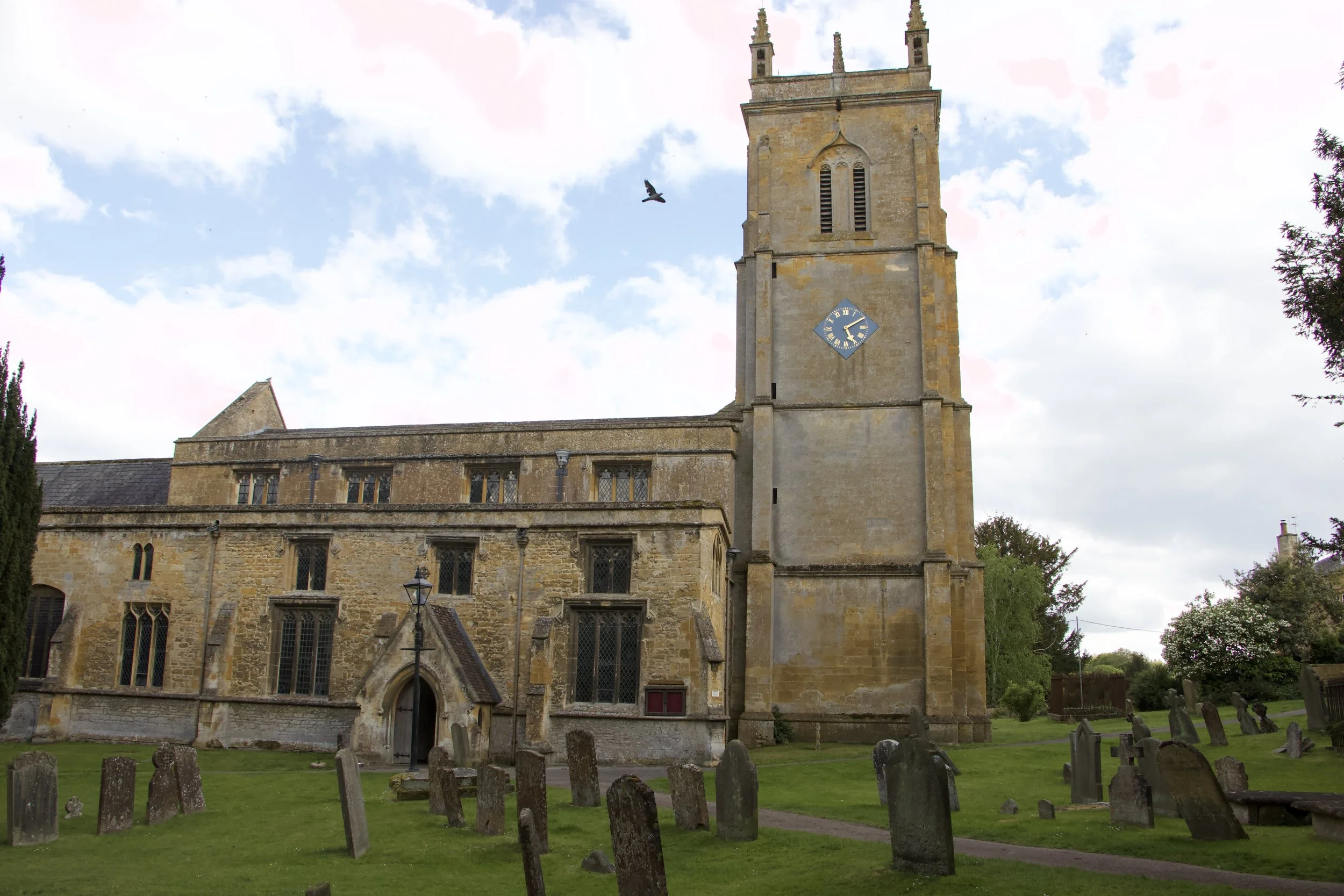 Historic stone English church, St. Peter and St. Paul's Church, with a clock tower, Gothic windows, and graveyard under a partly cloudy sky.