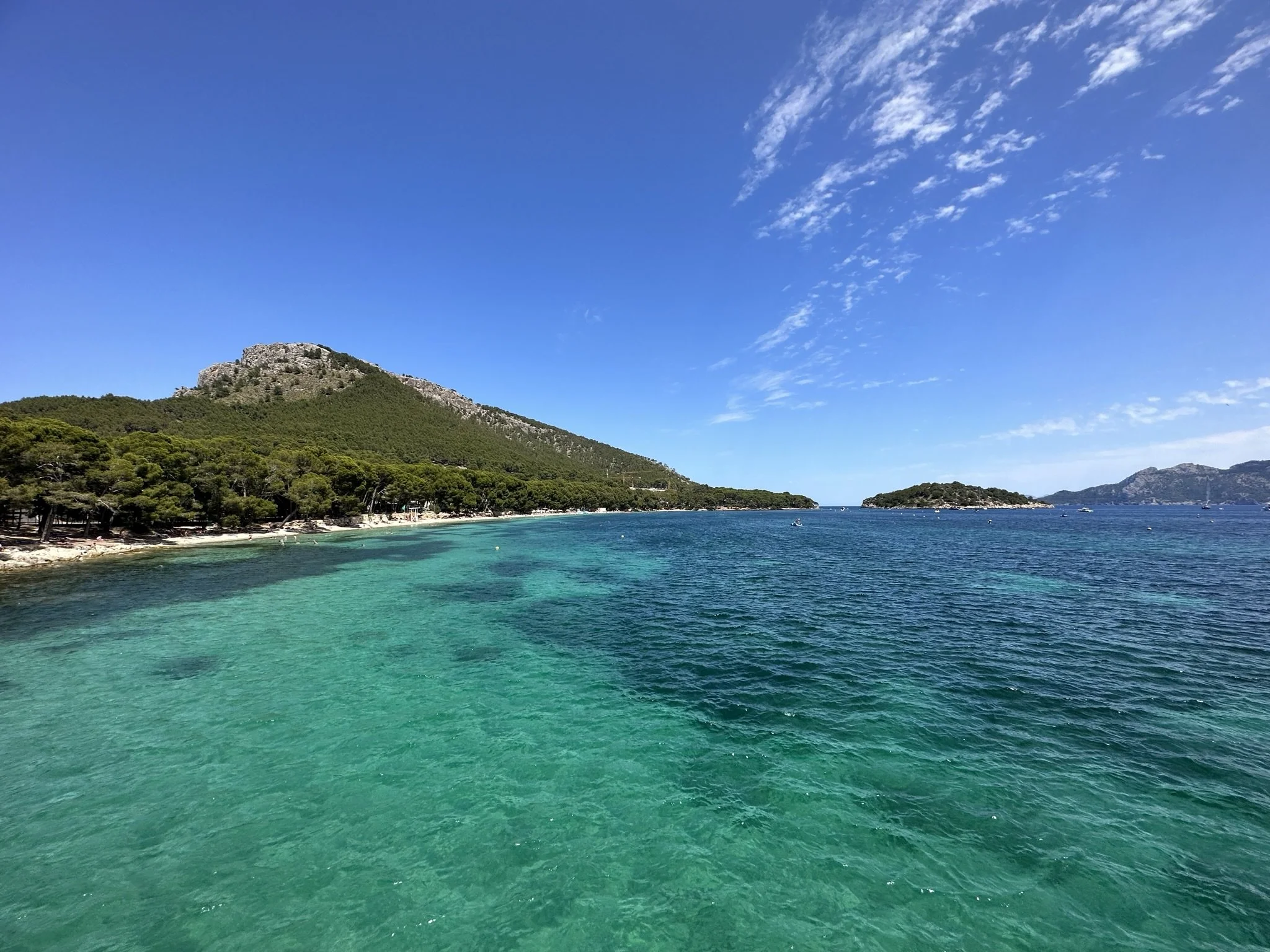 Turquoise Mediterranean sea and sandy beach beneath a forested rocky mountain on a sunny day