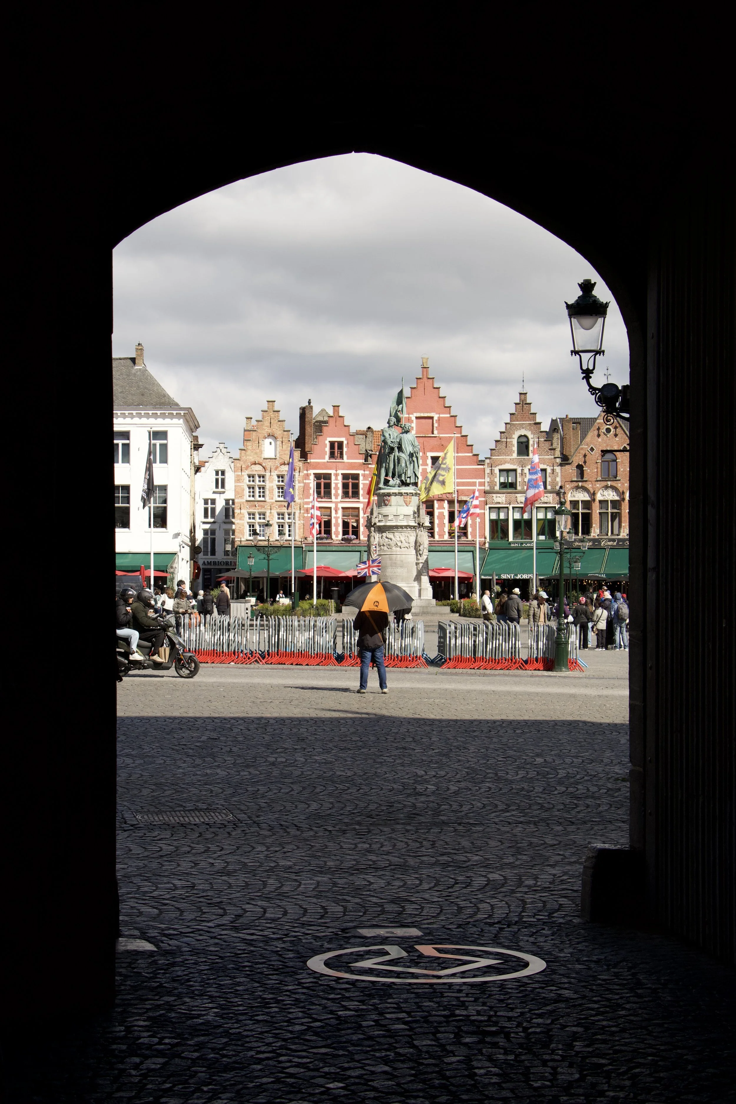 View of Bruges’ historic Markt square through a stone archway, featuring stepped-gable buildings, flags, a statue, cyclists, and cobblestone pavement.