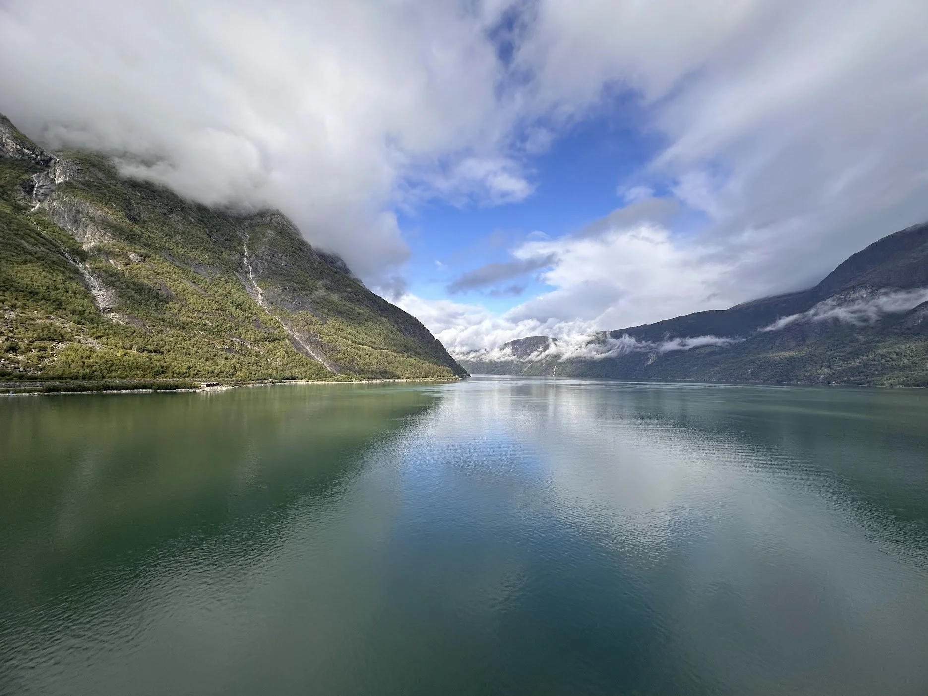 Scenic Norwegian fjord surrounded by steep, forested mountains, waterfalls, mist, and blue sky