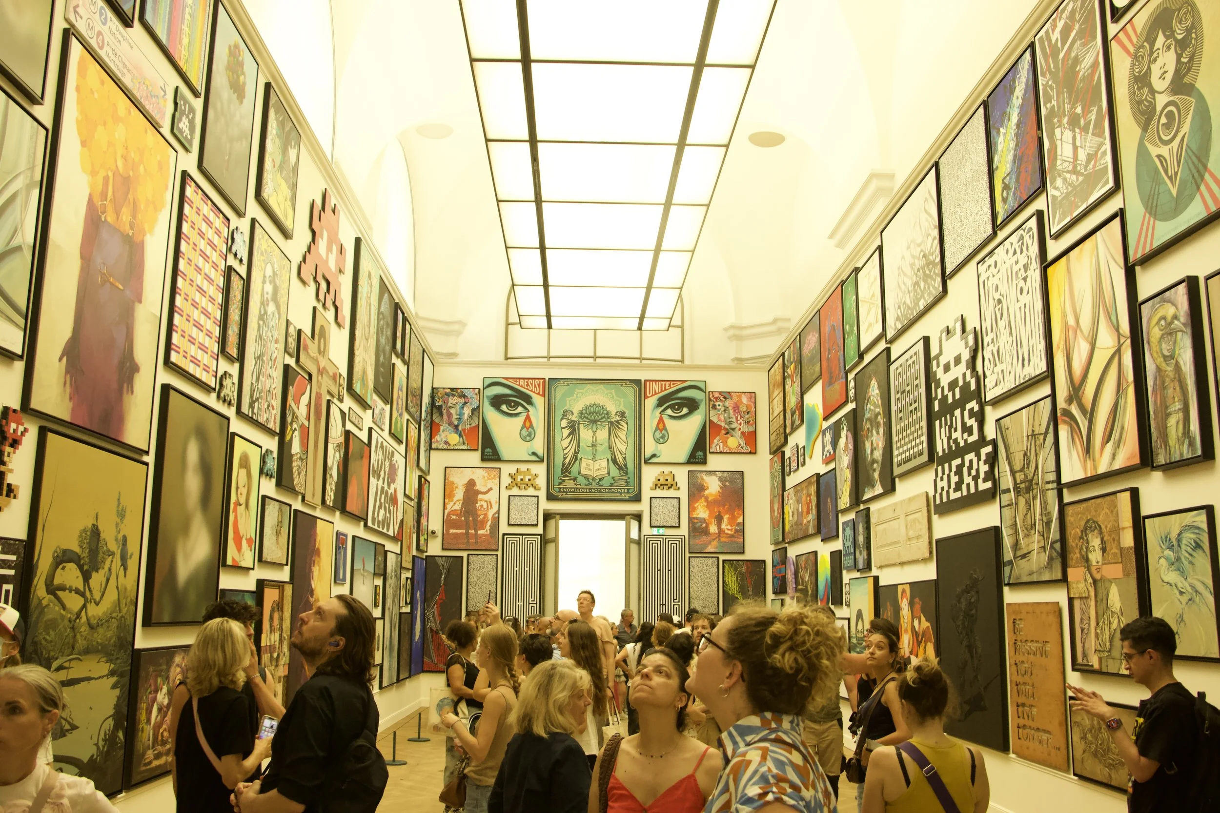 Crowded gallery hallway filled with framed contemporary artwork and visitors under a bright grid ceiling
