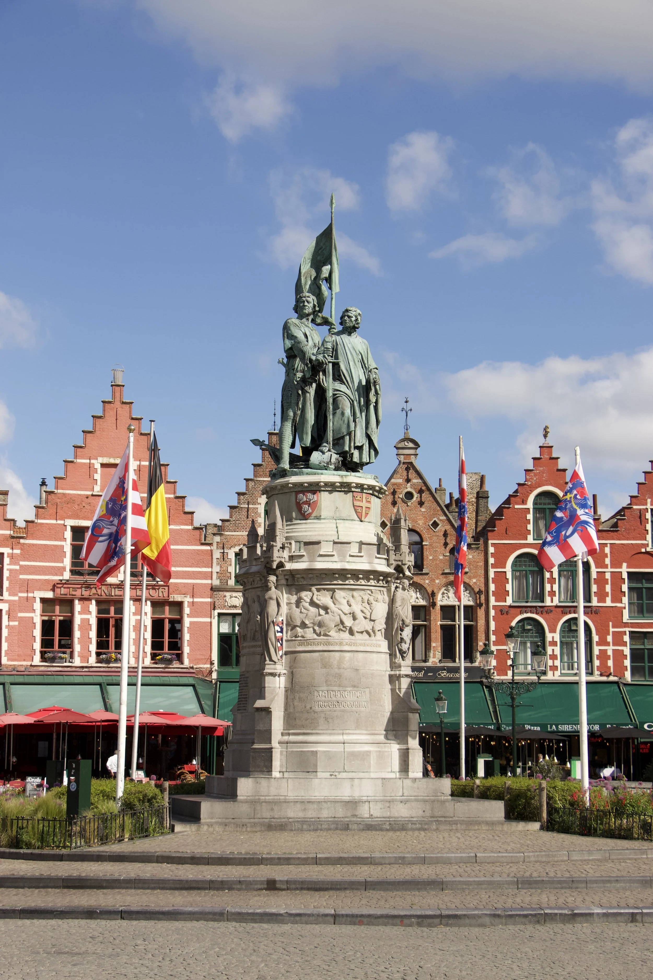 Bronze statue of Jan Breydel and Pieter de Coninck in Bruges’ Markt square, Belgium, with colorful flags and historic brick buildings.