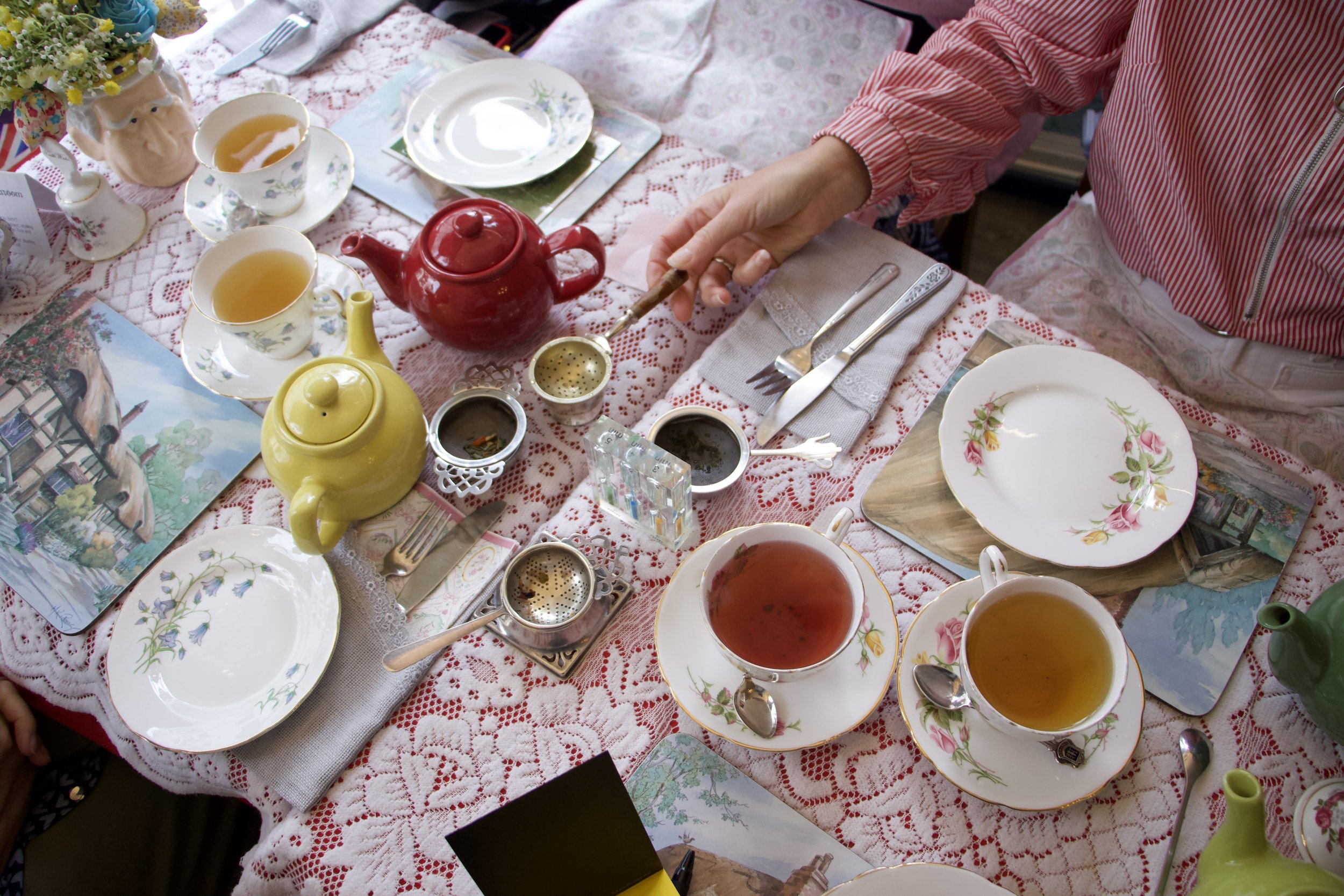 Vintage tea party table with colorful teapots, floral china, teacups, strainers, and a person holding a tea strainer at Leman Tea Room. 