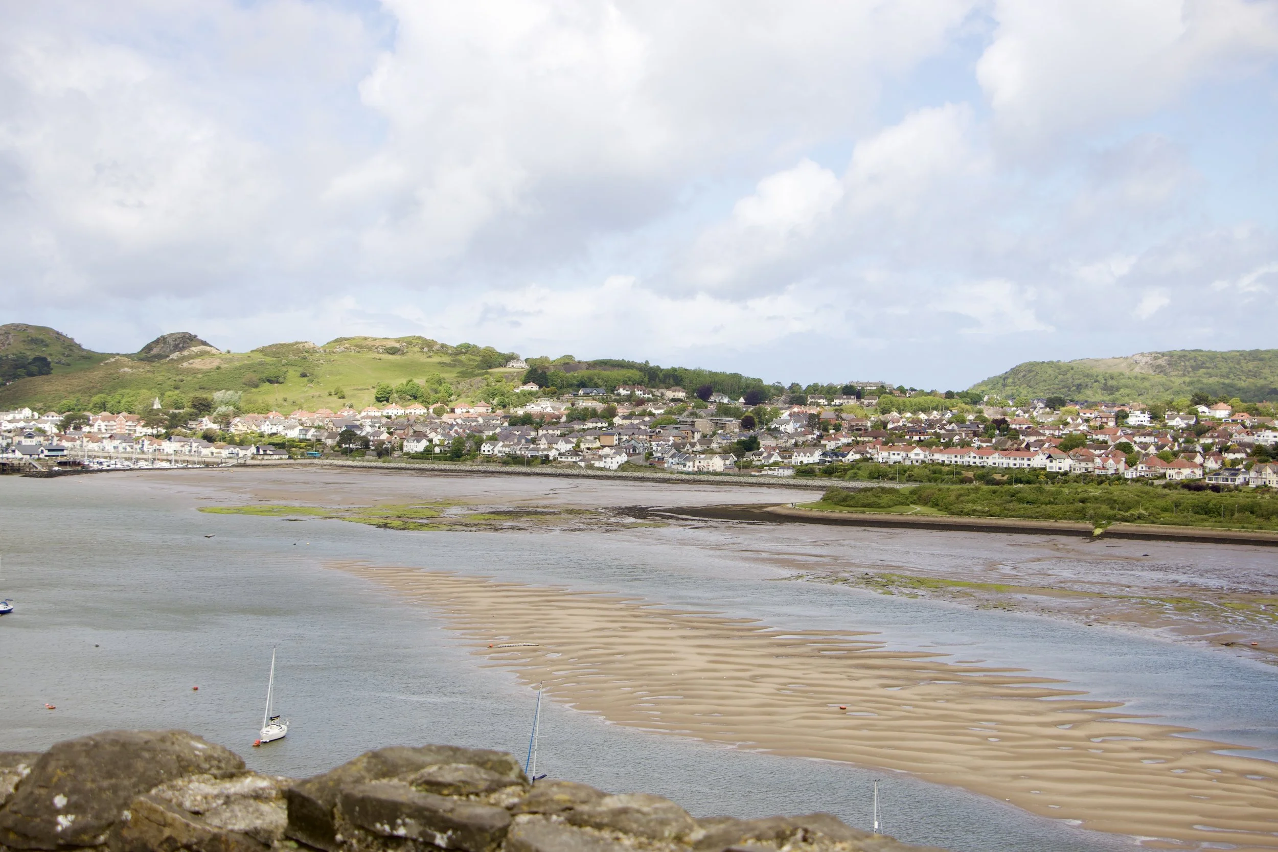 Panoramic coastal town with sailboats on tidal flats, rolling green hills, and cloudy skies.