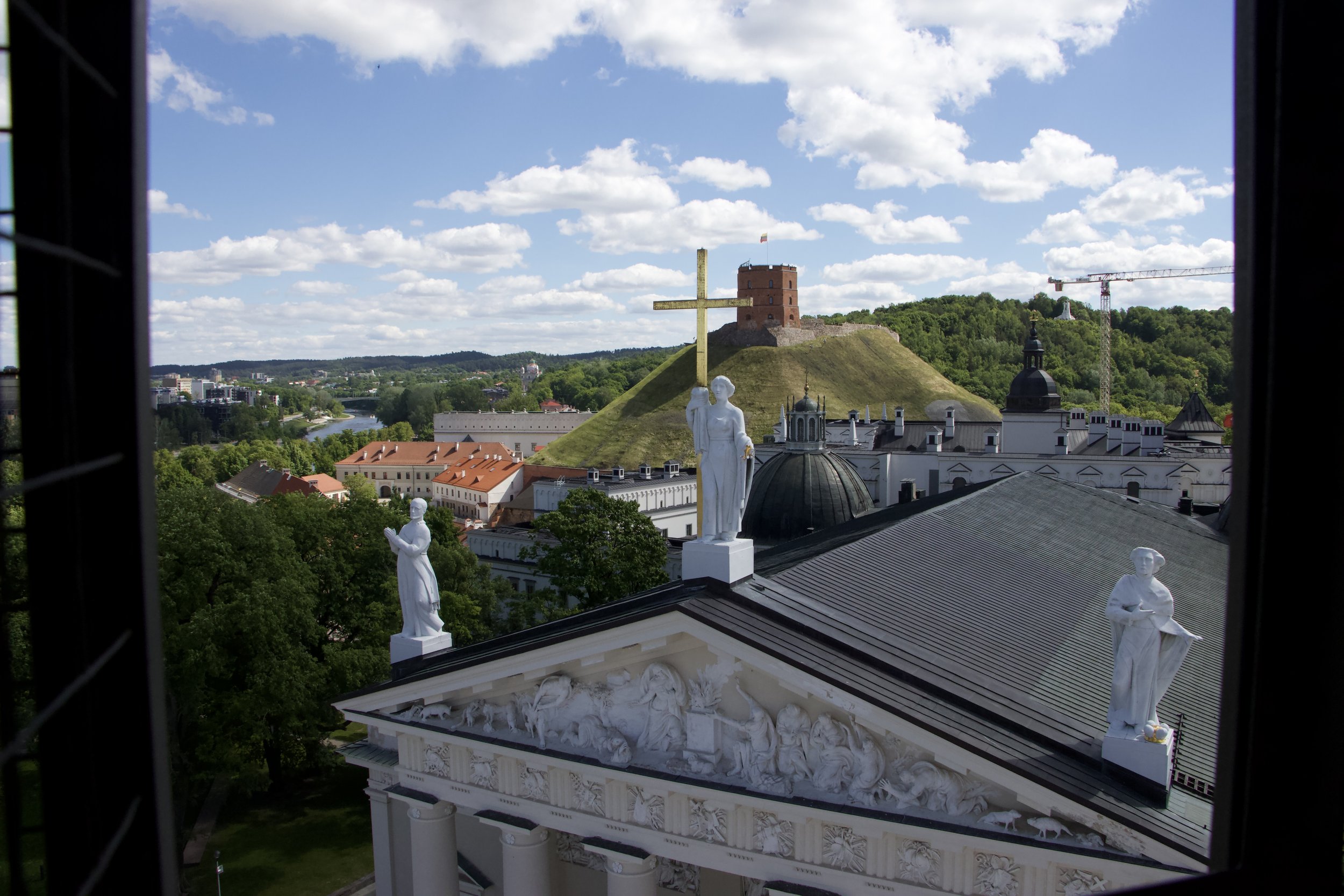 View of Vilnius, Lithuania, featuring Vilnius Cathedral statues, Gediminas Tower, and a large hilltop cross under a blue sky dotted with clouds.