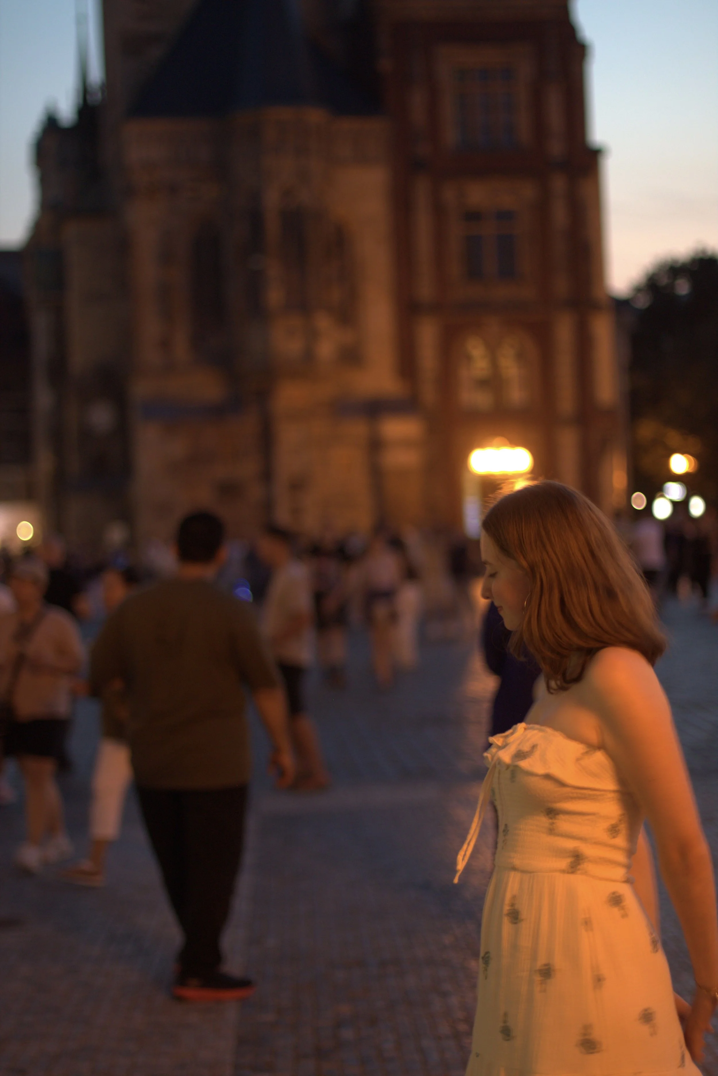 Blurry evening street scene with people walking past warmly lit historic brick buildings.