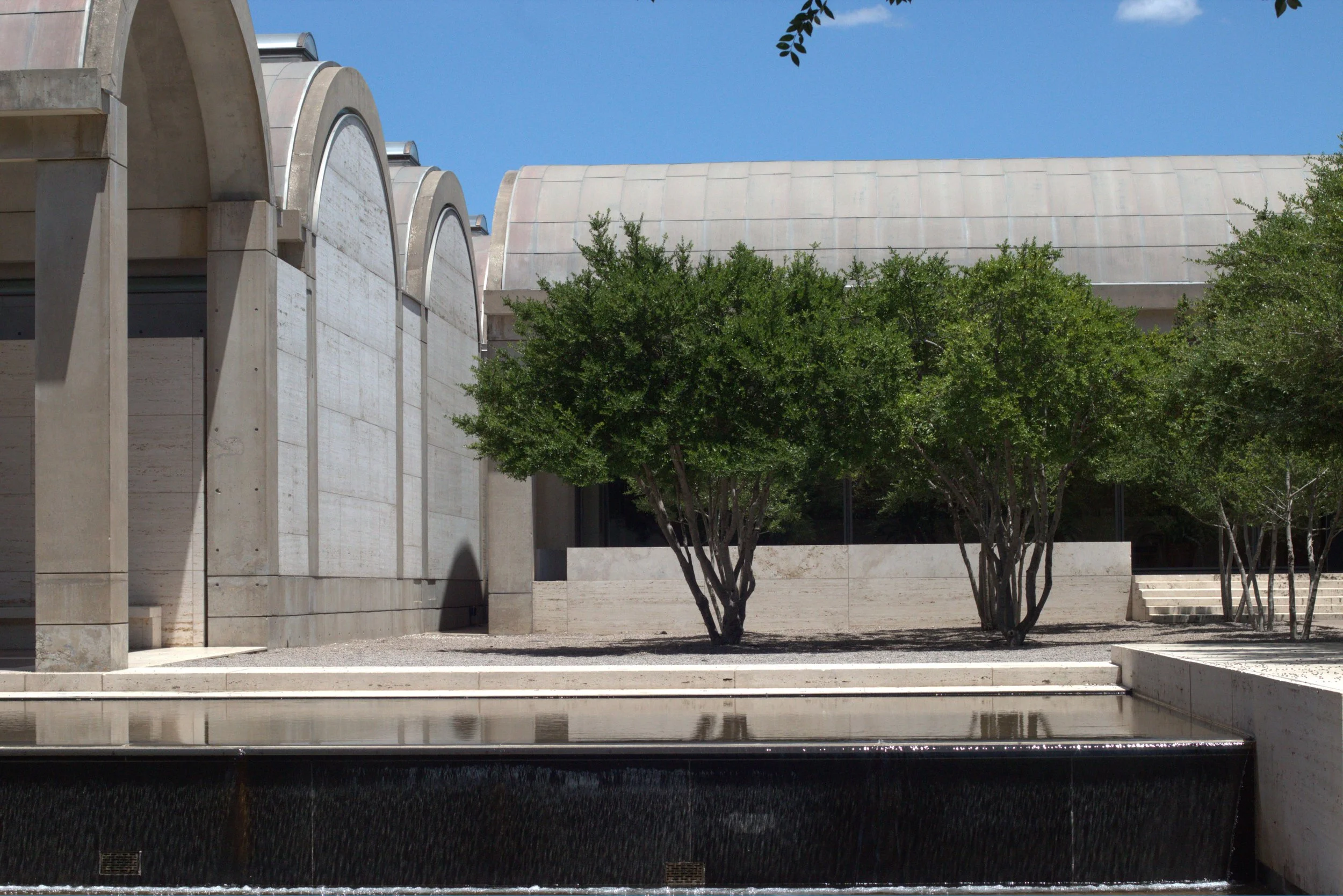 Modern concrete building with curved architectural walls, trees, and a reflecting pool under a clear blue sky