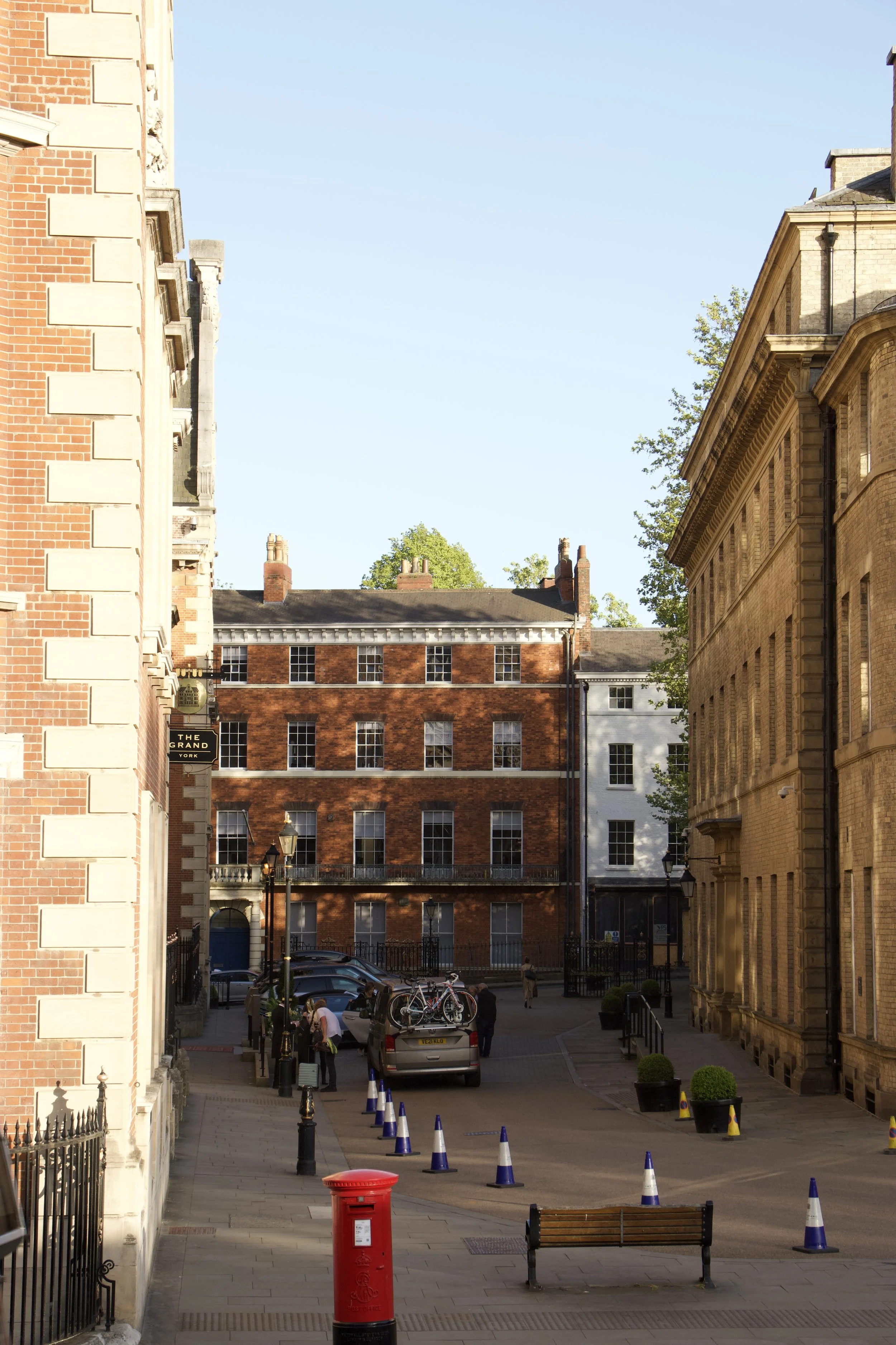 Historic York street with red-brick buildings, bicycle-topped car, traffic cones, benches, and a red British post box